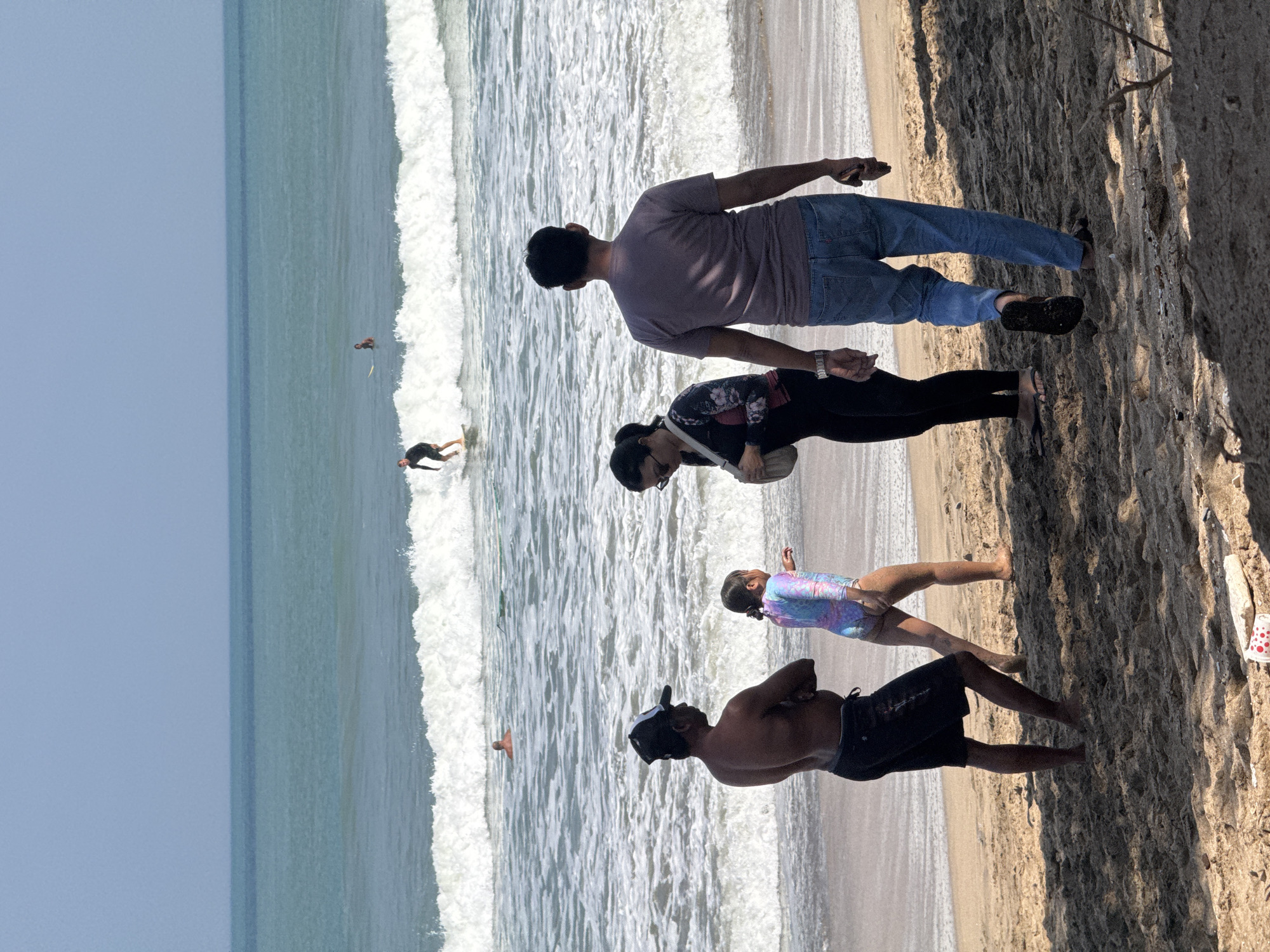 The image depicts a sunny day at a beach in Kuta, Indonesia, with a view of the ocean. In the foreground, two adults and a young girl are on the sand. A man wearing a black cap and dark shorts stands with his arms crossed, facing left. Next to him, a young girl in a colorful swimsuit is walking away from the camera towards the right. A woman wearing a patterned long-sleeved top and black pants, carrying a light-colored bag, walks towards the right, slightly behind the man and girl. Further to the right, a man in a purple t-shirt and blue jeans walks away from the camera.
In the middle ground, the ocean has breaking waves. Two surfers are visible in the water. One surfer, dressed in black, is riding a wave. Another surfer is further out in the water, and a third person is also visible in the water. The sky above is a clear, light blue. The sand in the foreground is light brown and textured. A discarded red and white object with red dots is visible on the sand in the lower left corner.