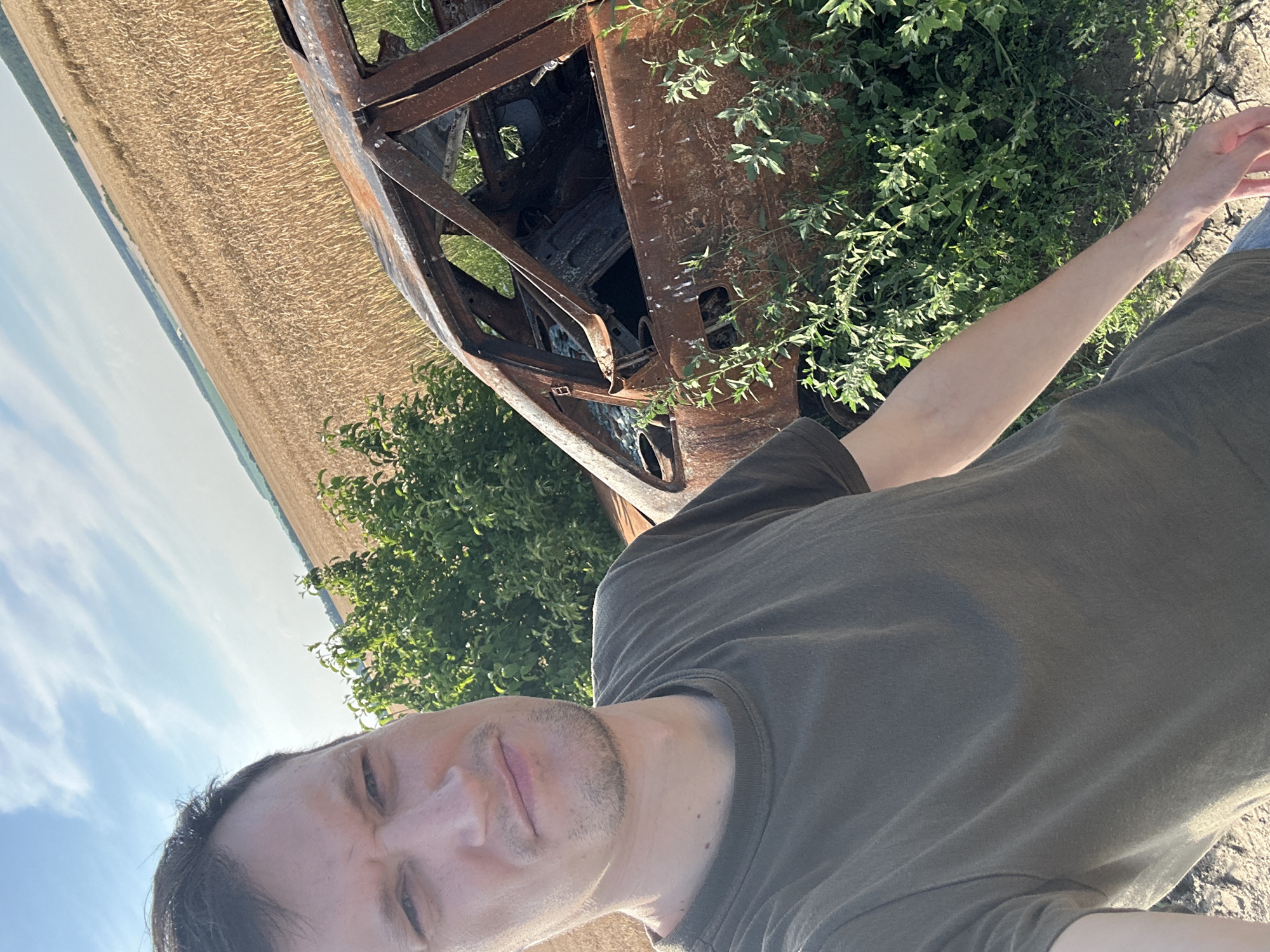 In Makiv, Ukraine, a man in a dark green t-shirt takes a selfie with a heavily rusted and burned-out car as the backdrop. The car appears to be a sedan, with its interior largely destroyed and exposed to the elements. Green vegetation grows around the vehicle, suggesting it has been there for some time. In the background, a vast field of golden wheat stretches towards the horizon under a bright blue sky with scattered clouds. The overall scene depicts a stark contrast between the man and the car, set against the tranquil beauty of the Ukrainian countryside.
