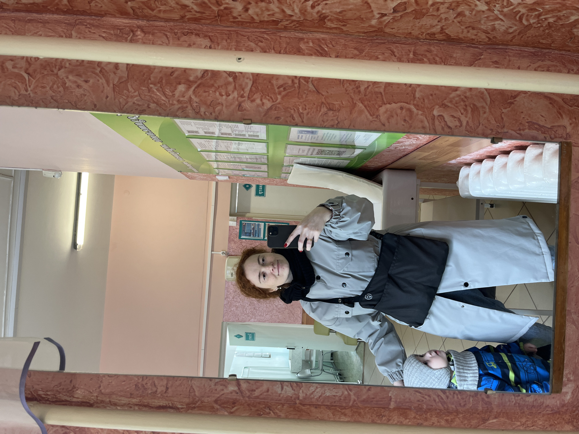 A selfie in a mirror shows a fair-skinned, red-haired woman wearing a light grey coat with a black scarf and black bag. She is holding a black smartphone to take the photo. In the reflection is a child lying down wearing a grey beanie and a blue and green striped snowsuit. The reflection also shows a clinical setting with information boards on the wall, medical equipment, and a doorway leading to another room with chairs. The photo was taken in the city of Chaikovsky, Russia.