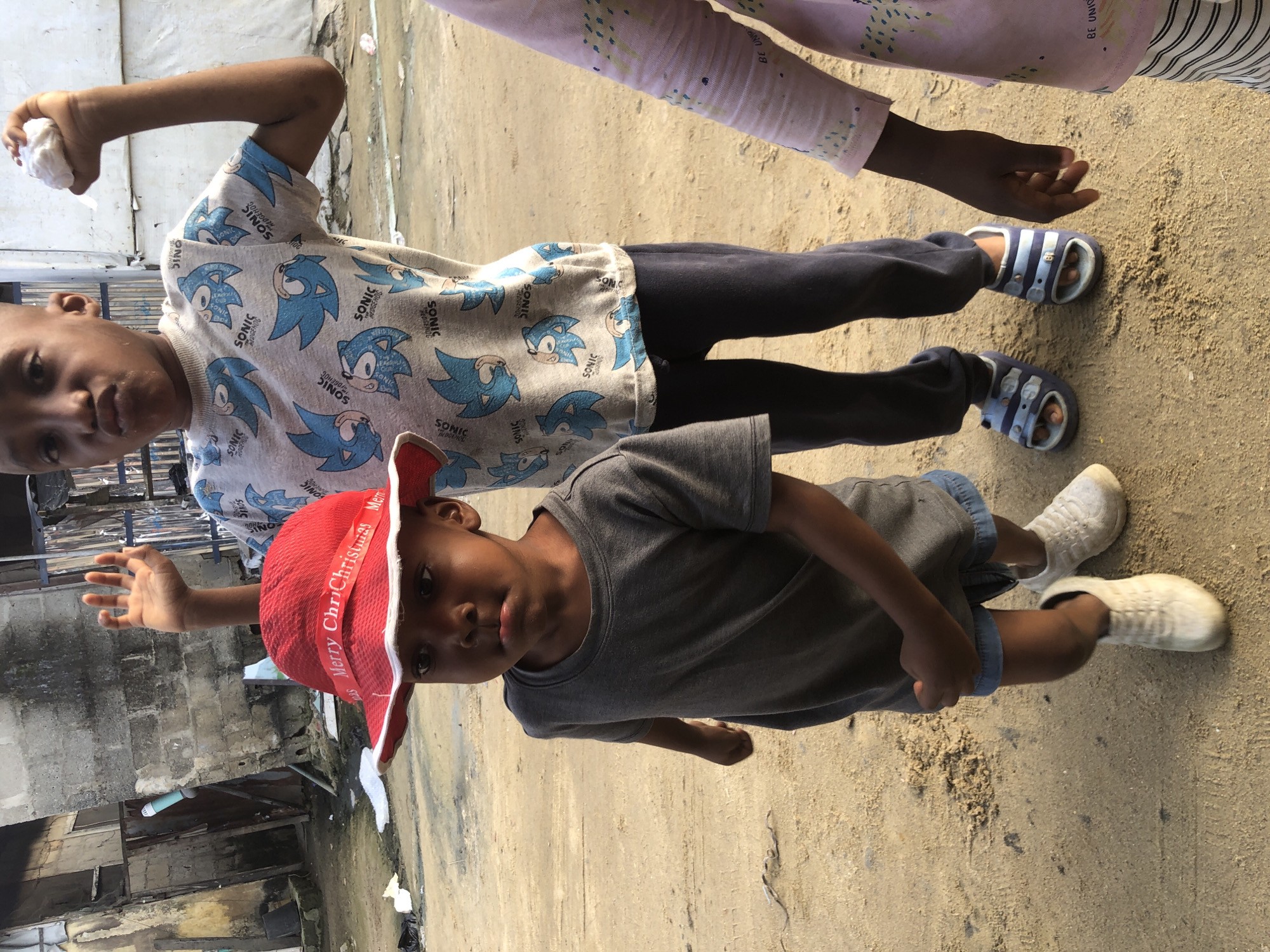 In Ado, Nigeria, a close-up shot captures a group of children in an outdoor setting, possibly a sandy or dirt path. The focus is on three individuals. In the foreground, a young boy wearing a gray t-shirt and blue denim shorts stands looking forward with a slightly pouting expression. He sports a red hat with "Merry Christmas" written on it in white lettering and white sneakers. To his right, an older boy, wearing a t-shirt adorned with Sonic the Hedgehog characters and dark pants, holds up a white object in his right hand and has his left hand raised. His gaze is directed towards the viewer. Partially visible on the right is a third child, wearing a light pink shirt with a patterned design. The background shows a dilapidated building with concrete walls and corrugated metal structures, suggesting an urban environment. The ground is sandy and shows some debris.
