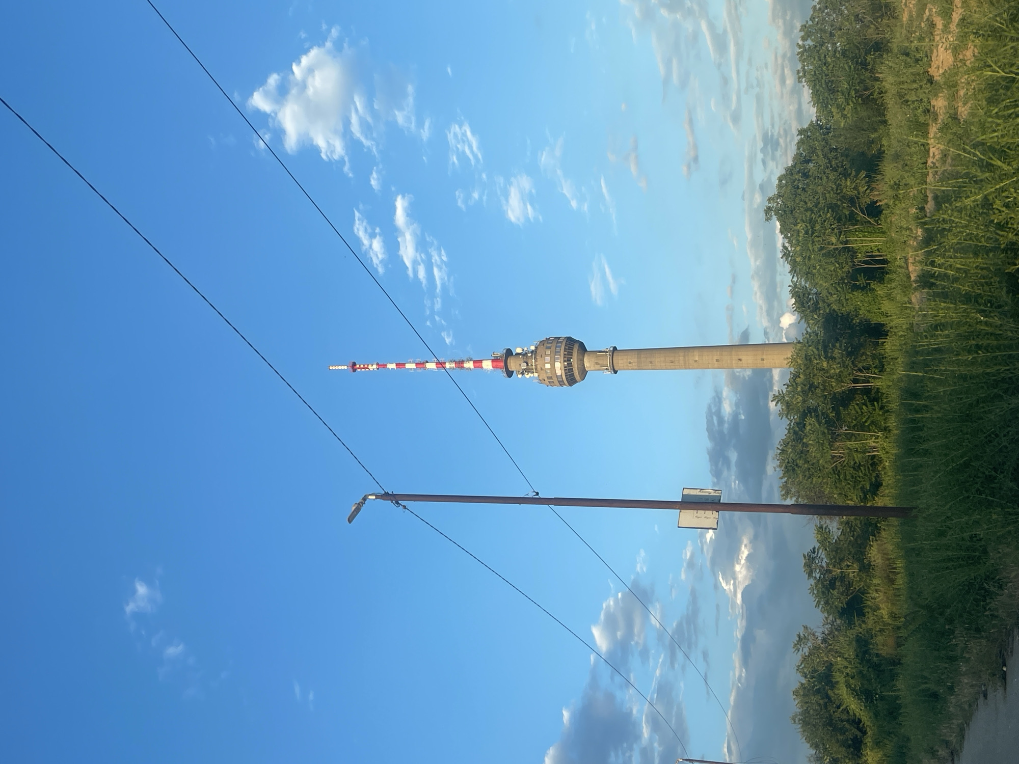 The image shows the Ruse TV Tower, also known as the Dunav TV Tower, located in Ruse, Bulgaria. The tower is a tall, concrete structure with a red and white striped antenna at the top. It is surrounded by lush green trees and vegetation. In the foreground, a lamppost stands tall, with power lines crisscrossing the frame. The sky is a vibrant blue, with scattered white clouds. The overall scene depicts a serene and picturesque view of the Ruse TV Tower against a beautiful natural backdrop.