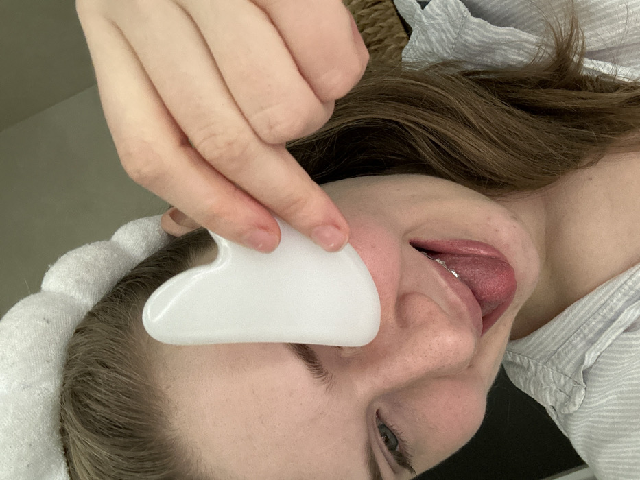 A woman is holding a white, heart-shaped tool against her face. She has a white, fluffy headband on, and her brown hair is partially visible. Her tongue is sticking out. Her eyes are open, and she is wearing braces. She is wearing a light-colored, striped shirt.