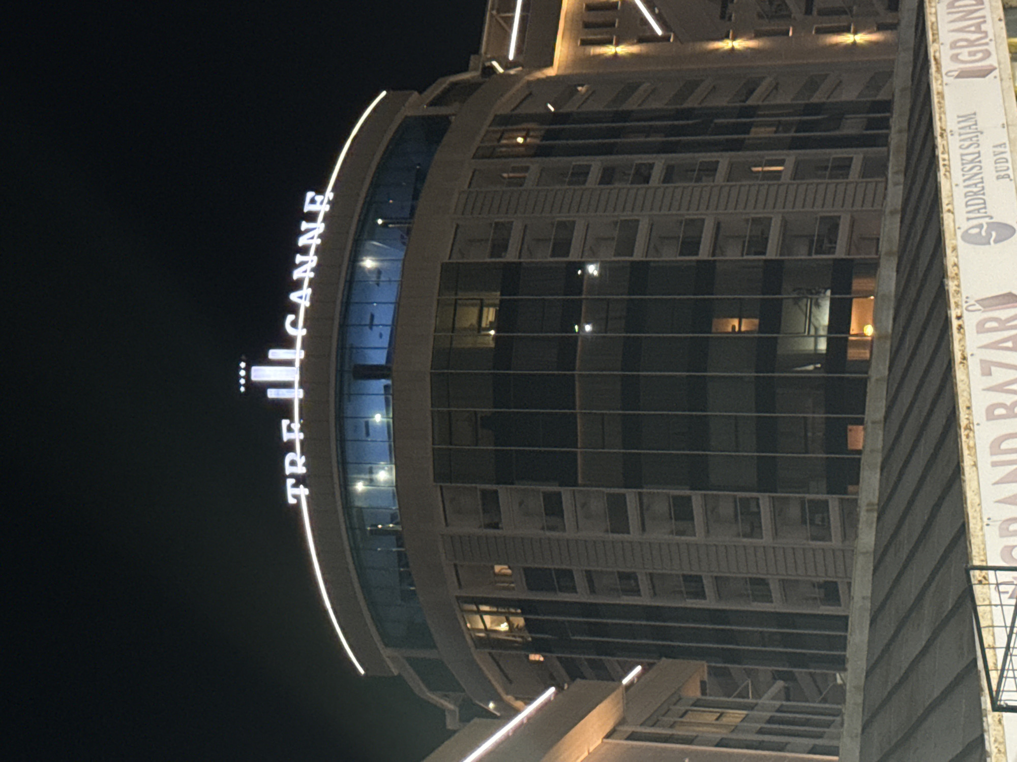 In the city of Budva, Montenegro, at night, a tall, modern building with a rounded facade is illuminated. The building has a curved glass-enclosed top floor with visible interior lighting. The main body of the building features numerous windows, some of which show interior lights, suggesting occupancy. A prominent neon sign in white letters, spelling out "TRE CANNE," is affixed to the top of the building. Below the main building, a dark, sloped roof structure is visible, and further down, a banner with text including "GRAND BAZAR" and "JADRANSKI SAJAM BUDVA" is partially visible. The scene is set against a dark night sky.