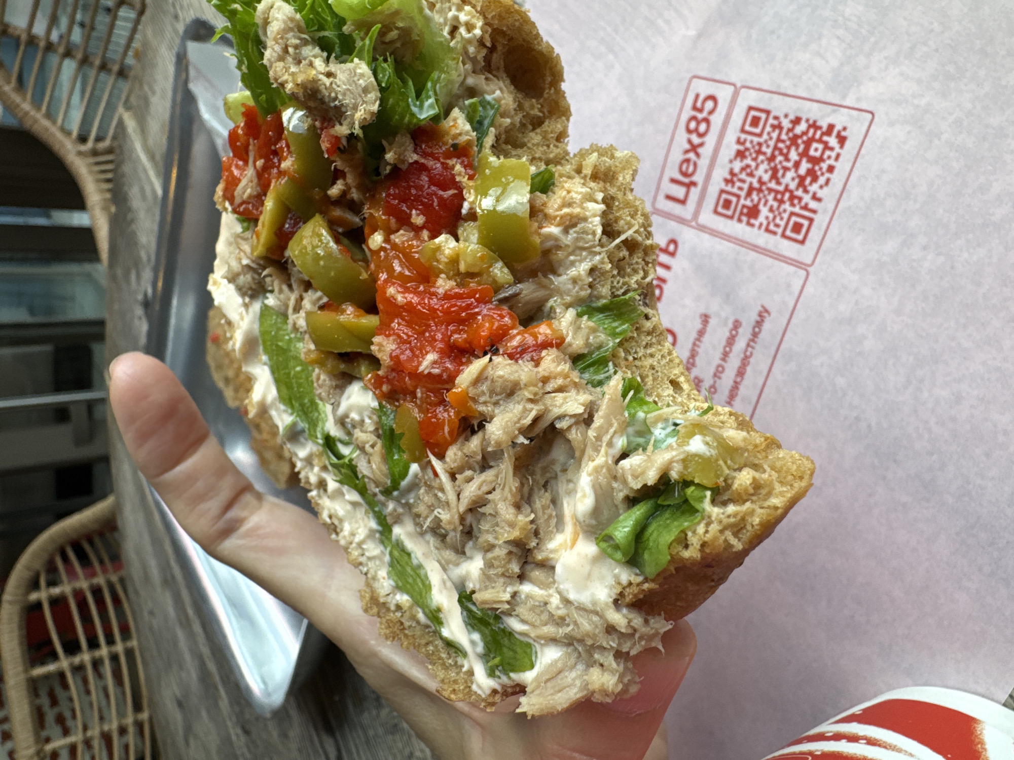 A person holds a half-eaten sandwich over a paper wrapper in what appears to be a cafe. The sandwich is made with whole wheat bread and is filled with shredded meat, lettuce, and what looks like chopped pickles and tomatoes. The wrapper has a QR code and text that reads "Цех85" which is the name of a bakery chain in Saint Petersburg, Russia. The background shows a wooden table and part of a wicker chair.