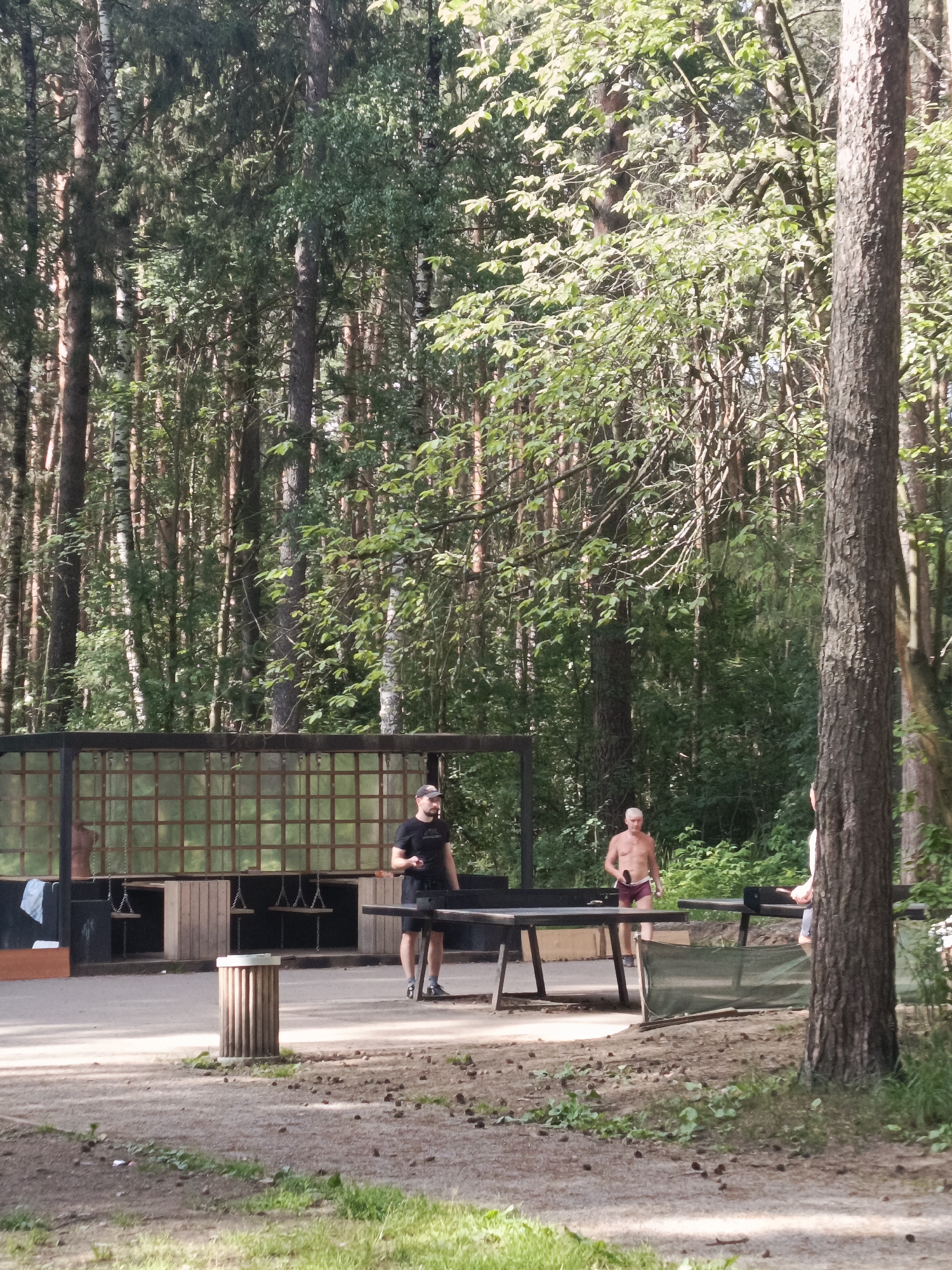 In an outdoor area with a forest backdrop, two men are engaged in a game of table tennis. The scene appears to be a designated sports or recreation zone, possibly within a park or recreational facility. The environment is characterized by dense trees, suggesting a natural or semi-natural setting. The ground is a mix of dirt and sparse vegetation.
The first man, on the left side of the frame, is wearing a black t-shirt and black shorts. He is standing on the left side of the table tennis table, holding a paddle in his right hand and a ball in his left hand, appearing to be in the process of serving or about to serve.
The second man, on the right side of the frame, is shirtless and wearing maroon shorts. He is positioned on the right side of the table, also holding a paddle, in a stance ready to receive a serve. His proximity to the table and paddle indicates active participation in the game.
A third individual, a woman, is partially visible on the far right of the image, with her back mostly to the camera. She is wearing a light-colored top and appears to be standing near the table tennis setup.
In the background, there is a wooden structure with a grid-like partition, possibly a pavilion or a covered seating area. Suspended from this structure are what appear to be swings, suggesting this area might also cater to other recreational activities. To the left of the table tennis table, a cylindrical trash receptacle is visible.
The overall impression is a casual, recreational activity taking place in a serene, tree-lined environment. The presence of multiple table tennis tables in the background hints at a larger facility for the sport. The image does not provide any explicit visual cues to confirm the location as the city of Perm, Russia.