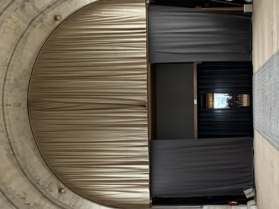 The image displays an interior architectural scene, with a large, light beige or tan pleated curtain dominating the left half of the frame. This curtain is arranged in a semi-circular arc, indicating it is part of a grand, possibly domed or rotunda-like structure. The surrounding wall, visible above and below the curved edge of the curtain, appears to be made of light-colored stone or concrete, featuring concentric arched bands that add to the sense of architectural grandeur. Two small, metallic light fixtures or attachment points are visible on this curved wall.
To the right of the beige curtain, the scene transitions into a more rectilinear area, characterized by darker curtains and structural elements. Two sets of dark grey or black curtains are drawn horizontally, creating a central opening. The upper dark curtain is pulled back to reveal an area beneath it, and similarly, the lower dark curtain is pulled back, revealing the space above it.
Within this central opening, a recessed area is visible, framed by dark wood or metal beams. Further back, another set of black curtains is partially parted, offering a glimpse into a deeper space. Behind these innermost curtains, an intricate, multi-layered structure of dark wood and possibly metal components is visible, suggestive of a pipe organ or a complex architectural element. Beyond this, a brightly illuminated, indistinct background can be seen.
On the dark wall to the left of this innermost opening, a small, square, illuminated white sign with dark text is present. The legible text on the sign shows the letter "A" followed by another character, possibly "C" or "G," stacked vertically. The flooring visible on the far right of the image consists of light-colored wooden planks, leading into a section of grey carpeting or a rug. A small, red object, possibly a fire extinguisher or safety device, is partially visible on the floor in the bottom right corner, near the dark grey curtain.
The overall setting suggests a formal interior space, such as a concert hall, theater, or a grand public building, possibly awaiting an event or performance. The lighting is artificial and uniform, indicating an indoor environment.