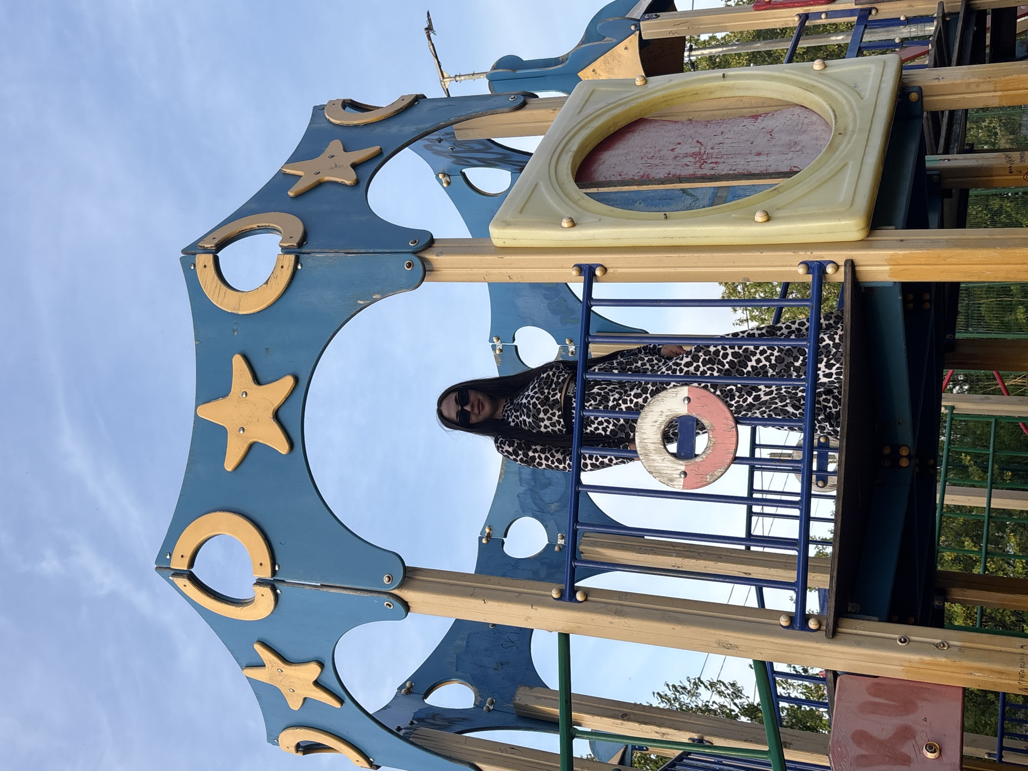 A medium shot captures a woman standing within a children's playground structure, likely in the city of Фоки, Russia. The woman is positioned inside the blue metal structure that is accented with gold stars and horseshoes, which forms the apparent exterior of a play tower. She is wearing dark sunglasses and a dress with a black and white pattern, and she appears to be looking directly at the camera. The playground structure features a combination of blue metal bars, yellow wood planks, and a beige square window, typical of playground equipment. The background shows more of the playground equipment, some trees, and a blue sky partially covered with clouds.