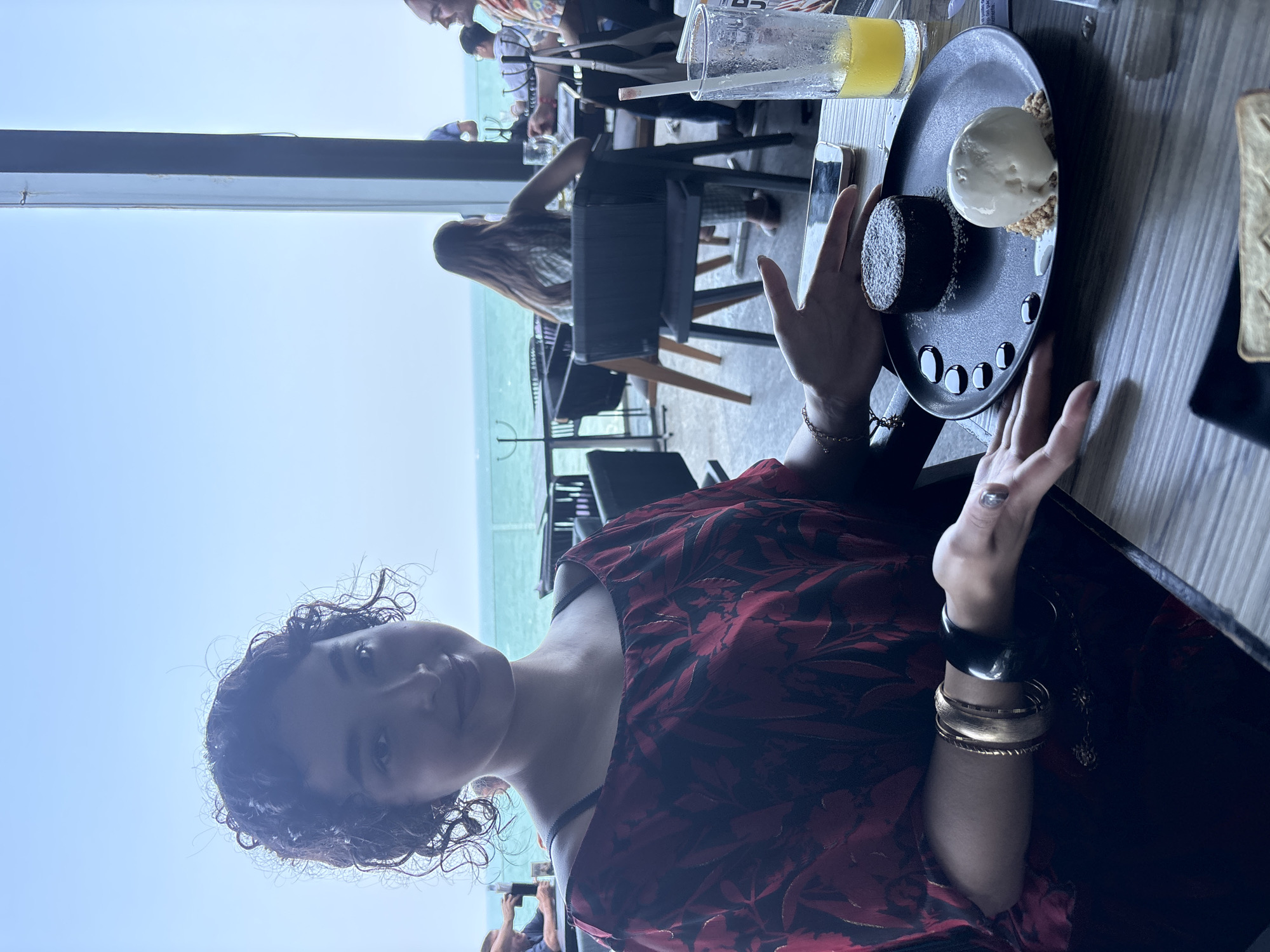 A young woman sits at a table in an outdoor restaurant, likely in Veracruz, Mexico, given the ocean view. She wears a red floral dress and several bracelets on her left wrist. In front of her, on a black plate, are a chocolate lava cake, a scoop of vanilla ice cream, and a series of small chocolate dots as decoration. A glass of orange juice and a cell phone are also on the table. In the background, other diners are seated at tables, and the ocean and sky are visible. The photo is taken from a slightly low angle, making the woman the primary focus.