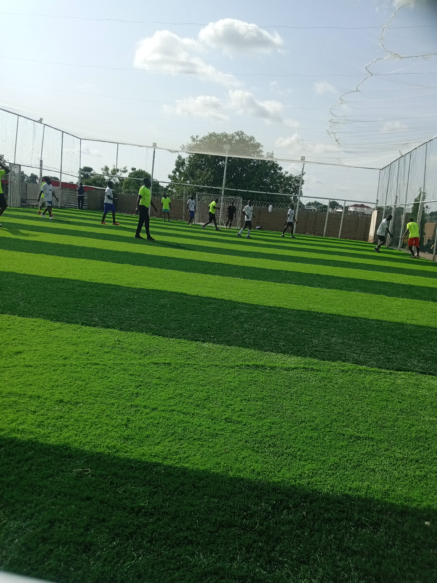 The image depicts an outdoor artificial turf football field in Jimeta, Nigeria. A group of young men, dressed in athletic wear, are engaged in a game of football. Some are actively playing, while others are observing or moving around the field. The field is enclosed by a fence and netting, suggesting it is a dedicated sports facility. The sky above is partly cloudy, and the time of day appears to be afternoon, judging by the shadows cast on the turf. The surrounding environment is characteristic of a developing urban area, with trees and some buildings visible beyond the fence.