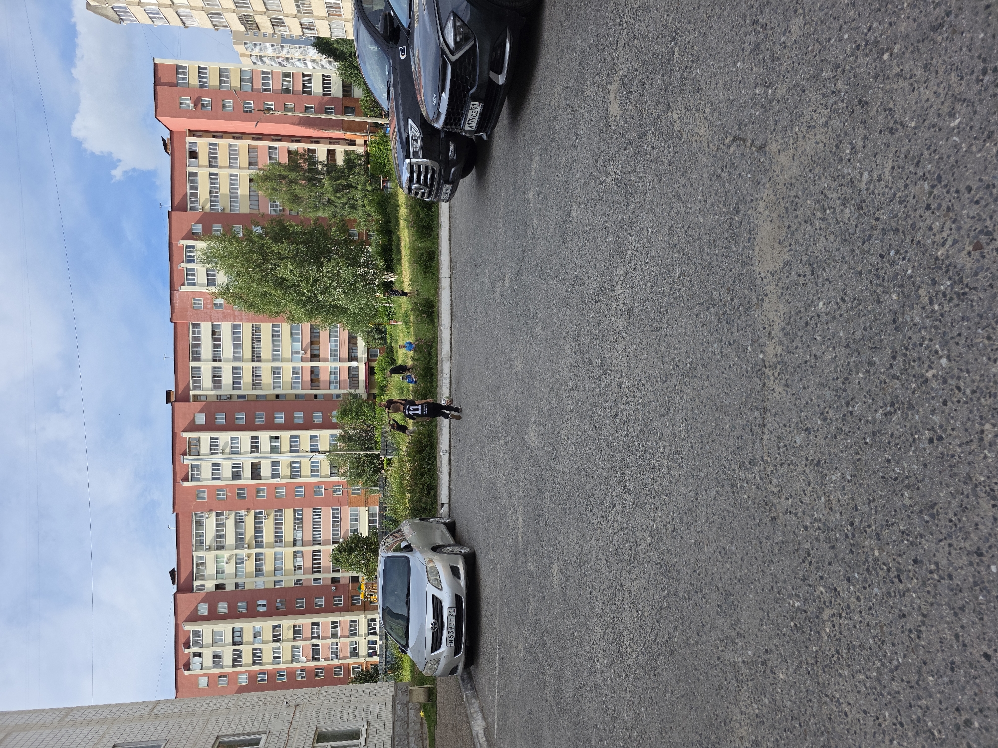 The image depicts a residential courtyard scene in Novocheboksarsk, Russia. In the foreground, there is an asphalt parking lot with several cars. A silver sedan, identified by the license plate H639ET 21, is parked on the left. To its right are two SUVs, a dark one with a partial license plate H260MX 21, and a dark gray one with the license plate H179CE 31.
In the middle ground, a patch of green grass and trees separates the parking lot from a large, multi-story residential building. The building is a prominent pink and yellow structure with numerous windows and balconies. Several individuals are visible in the grassy area, including a person wearing a black shirt with the number 11 on the back, and a few other figures who appear to be children or young adults.
The background is dominated by the apartment building and a sky with scattered clouds. The overall impression is a typical urban residential environment.