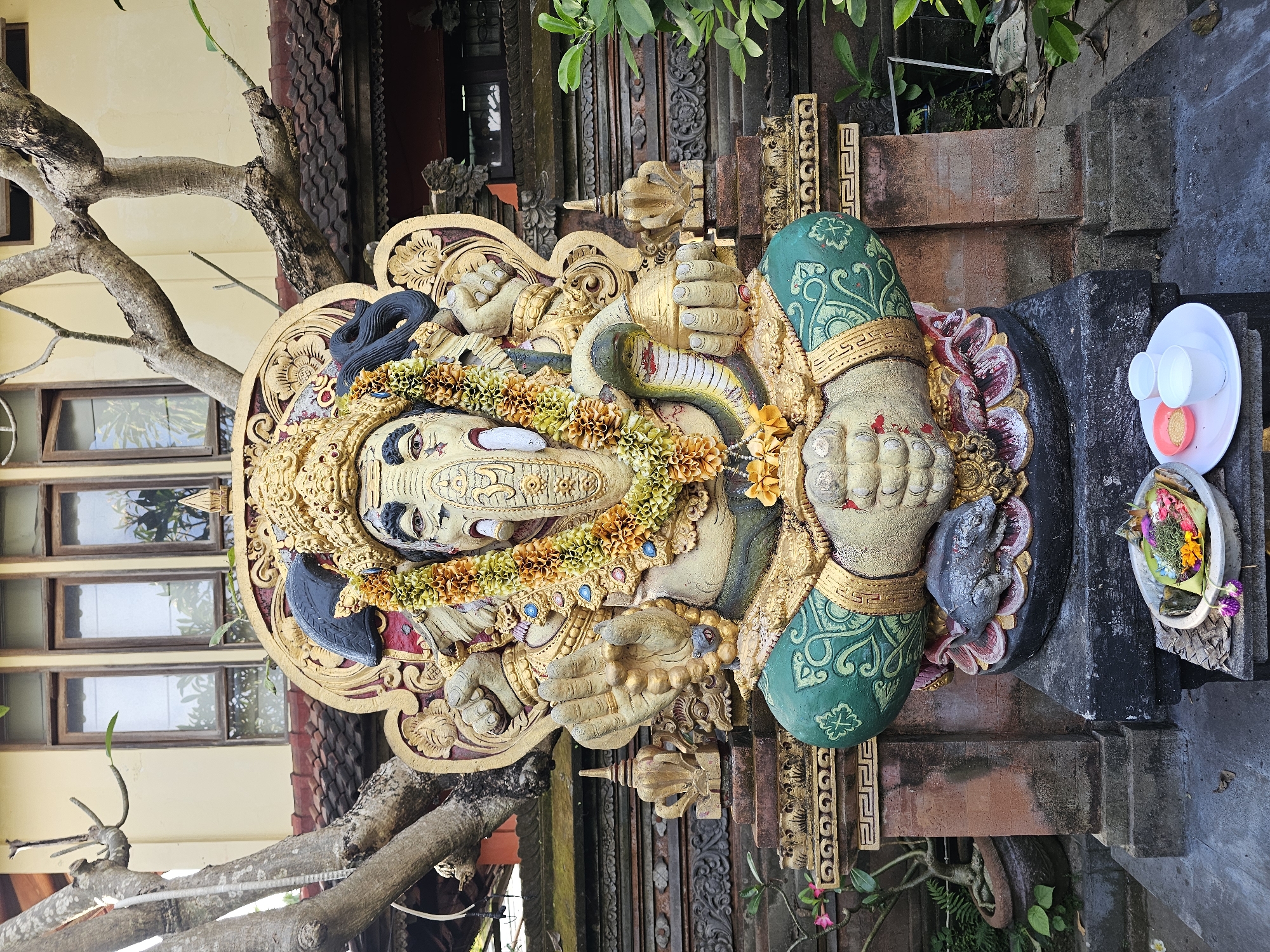 The image shows a statue of Ganesha, the Hindu god of wisdom and prosperity, located in Denpasar, Indonesia. The statue is elaborately decorated with a garland of yellow and orange flowers, and features intricate carvings and gold detailing. The statue has a snake wrapped across its torso. The statue is set against a background of Balinese architecture and foliage. To the right of the statue is a white plate with offerings.