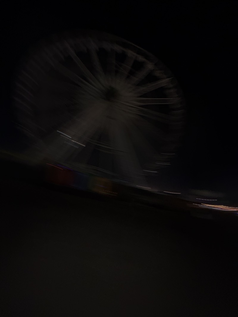 A blurred photograph depicts a Ferris wheel at night, likely in motion. Light sources create streaks, suggesting a slow shutter speed or movement during the exposure. The lower portion of the image is predominantly dark. The location of the image cannot be determined from the image.
