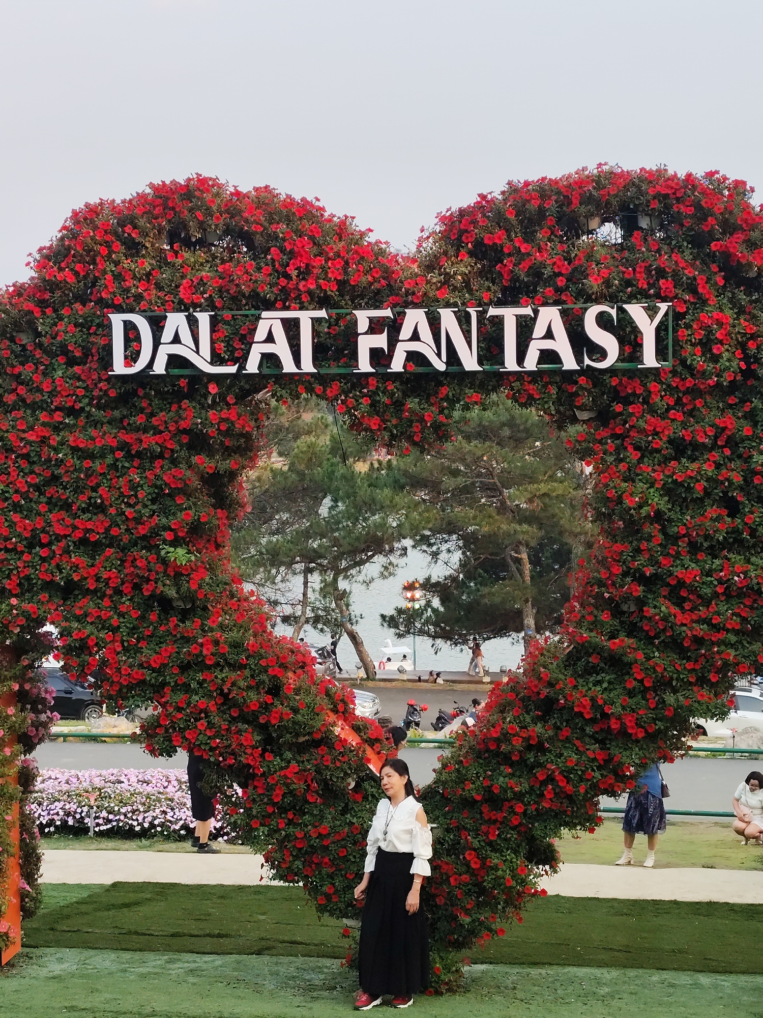 A full outdoor medium shot presents a heart-shaped floral structure with the words "DALAT FANTASY" in white lettering across the top. The heart is made of small red flowers. A woman with dark hair, dressed in a white blouse and black skirt with white-and-red shoes, stands on the right side of the heart. She faces forward and looks slightly to the left. In the background, through the heart-shaped opening, there is a glimpse of a body of water, trees, and distant figures. To the left of the heart structure, there is a garden area with pink flowers. The ground near the heart structure is green, possibly grass or artificial turf. Other people are visible in the background, including some near the water and others on a pathway. Cars and motorcycles are parked along the road. The scene is likely in Da Lat, Vietnam, given the lettering on the floral structure.