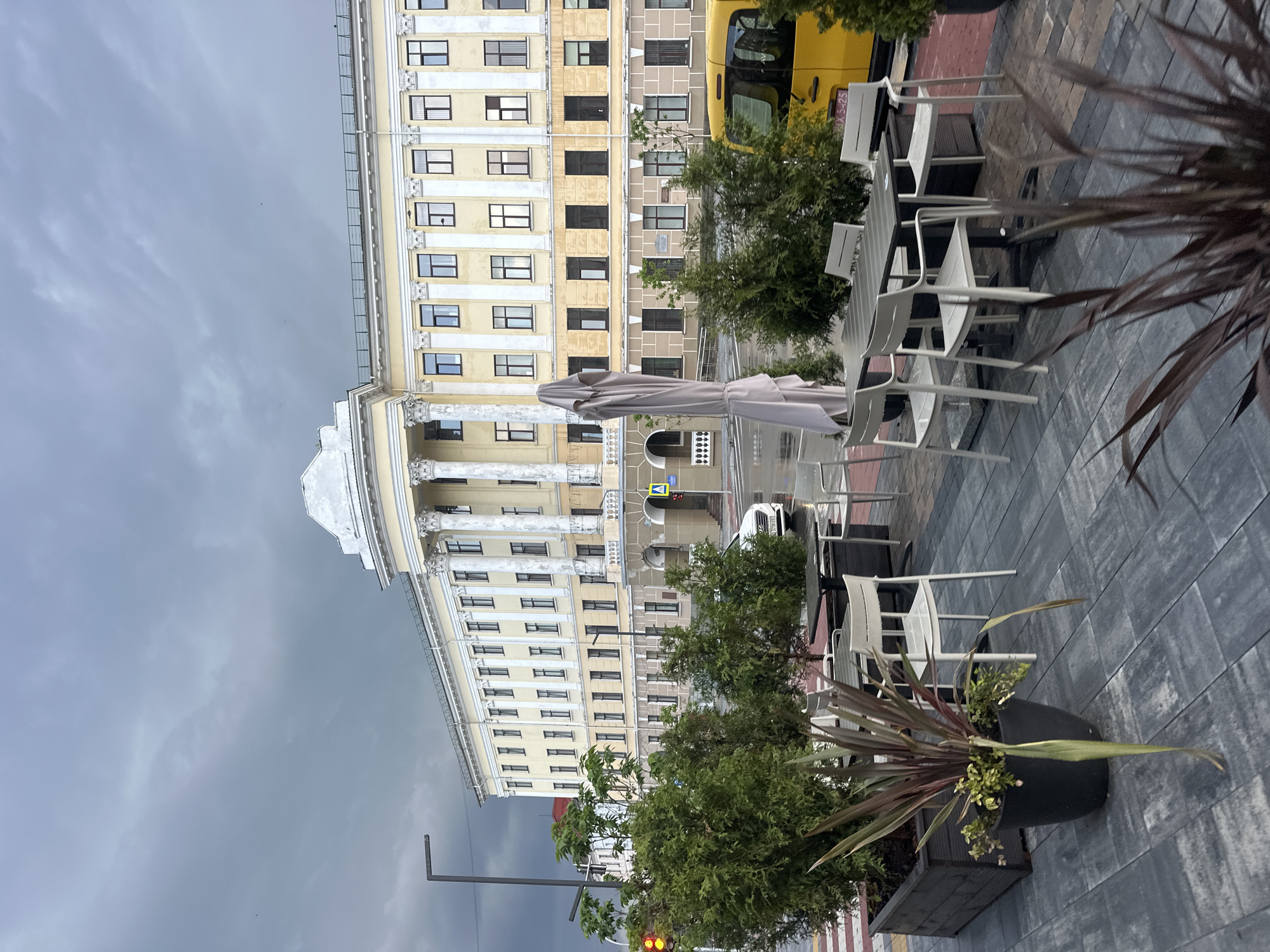 An outdoor seating area with tables and chairs is in the foreground, with a large building in the background. The building is light yellow with white columns and has many windows. A yellow van is parked on the street to the right of the building. The sky is cloudy and gray. The scene is likely from Ternopil, Ukraine, given the architecture and the presence of a yellow van, which is a common type of vehicle used for public transportation in Ukraine. The area appears to be a public space, possibly a cafe or restaurant.