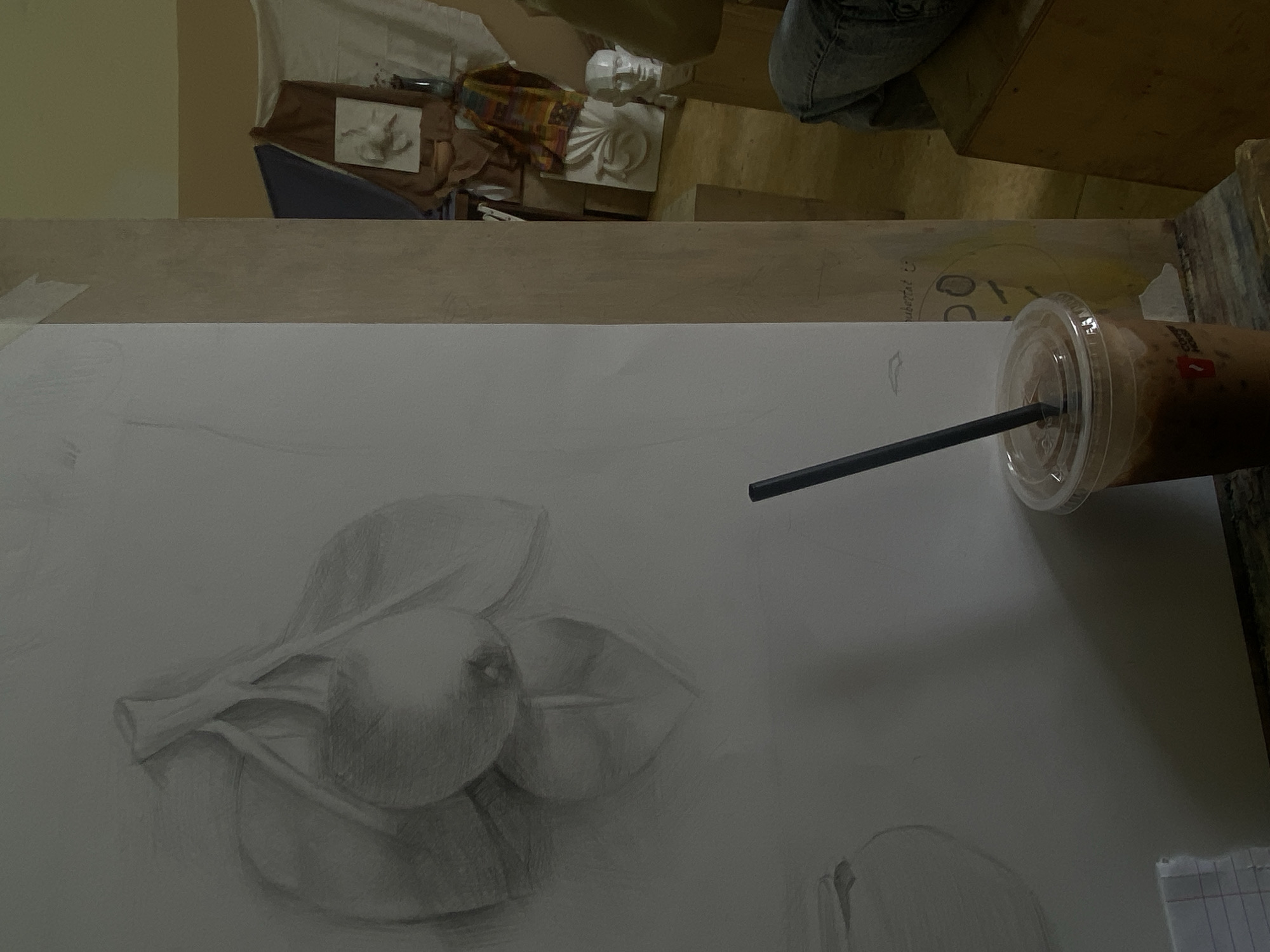 The image depicts a close-up shot of an artist's workspace. The primary subject is a drawing in progress on a large sheet of white paper, featuring a still life composition of fruits, likely apples or pears, with leaves. The drawing is rendered in a graphite pencil style with shading.
A plastic cup with a lid and a black straw sits to the right of the drawing, indicating a beverage.
The workspace appears to be on a table or desk, with part of a wooden frame visible on the right side.
In the background, out of focus, are other art supplies and a mannequin, suggesting an art studio setting. The location of Yerevan, Armenia, cannot be verified from the visual information provided.