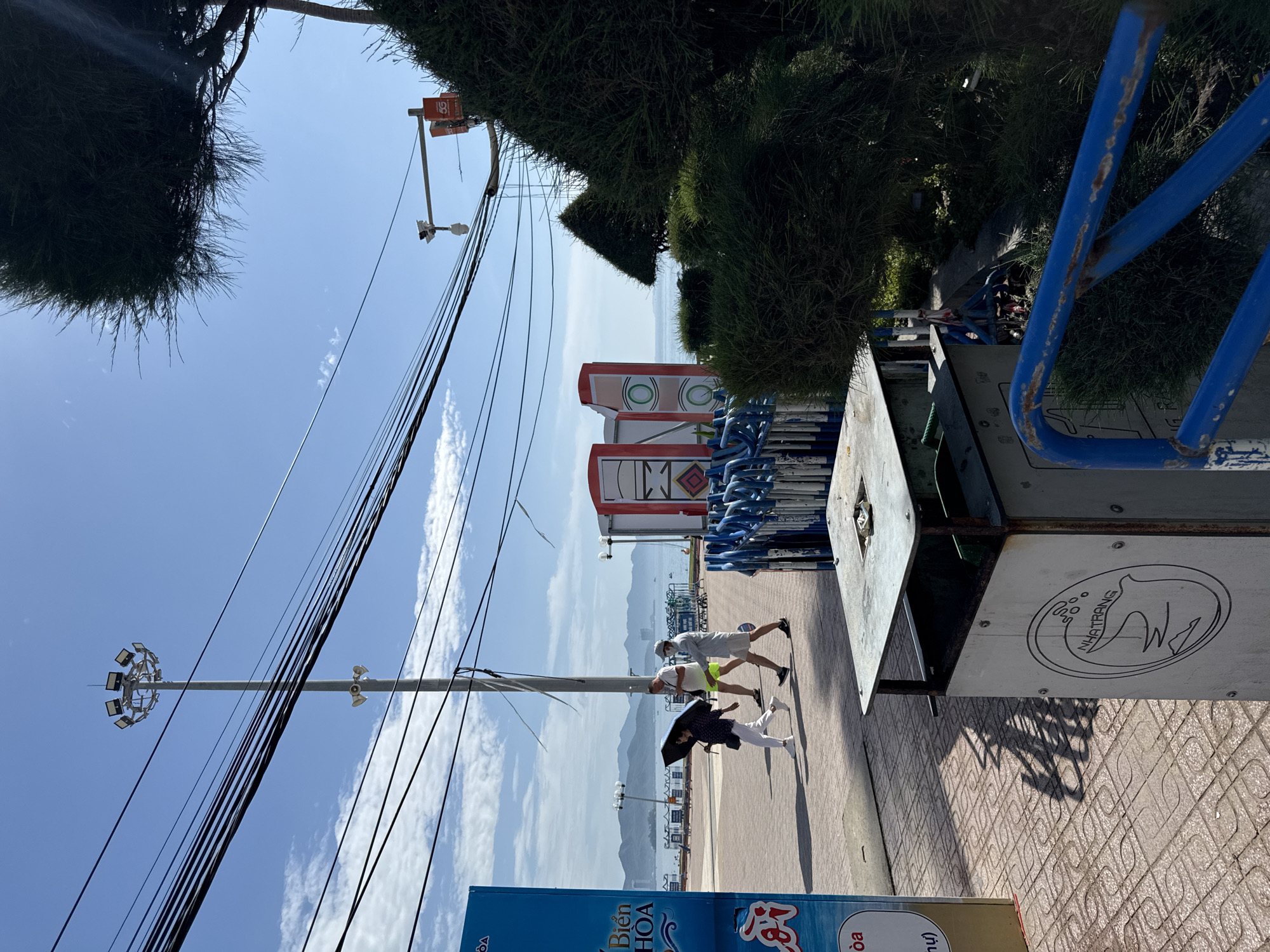 The image shows a sunny day in Nha Trang, Vietnam. In the foreground, a metal trash receptacle with a circular emblem depicting a bird and the word "NHATRANG" is visible on the right. To the left of the trash receptacle, a section of a blue barrier is present. The ground is paved with reddish-brown tiles laid in a grid pattern, with shadows cast by unseen objects. Three people are walking on the tiled pathway. The person on the left is holding a black umbrella and is wearing white pants and a patterned top. The middle person is a male wearing bright yellow shorts and a dark t-shirt. The person on the right is wearing white shorts and a light-colored shirt, with a white face mask. Behind them, the sea stretches to the horizon, with mountains visible in the distance under a clear blue sky. Several power lines stretch across the frame, and a tall light pole with multiple lamps is prominent. Two red and white structures with circular green designs are situated between the people and the sea. A dense green tree partially obscures the right side of the image.