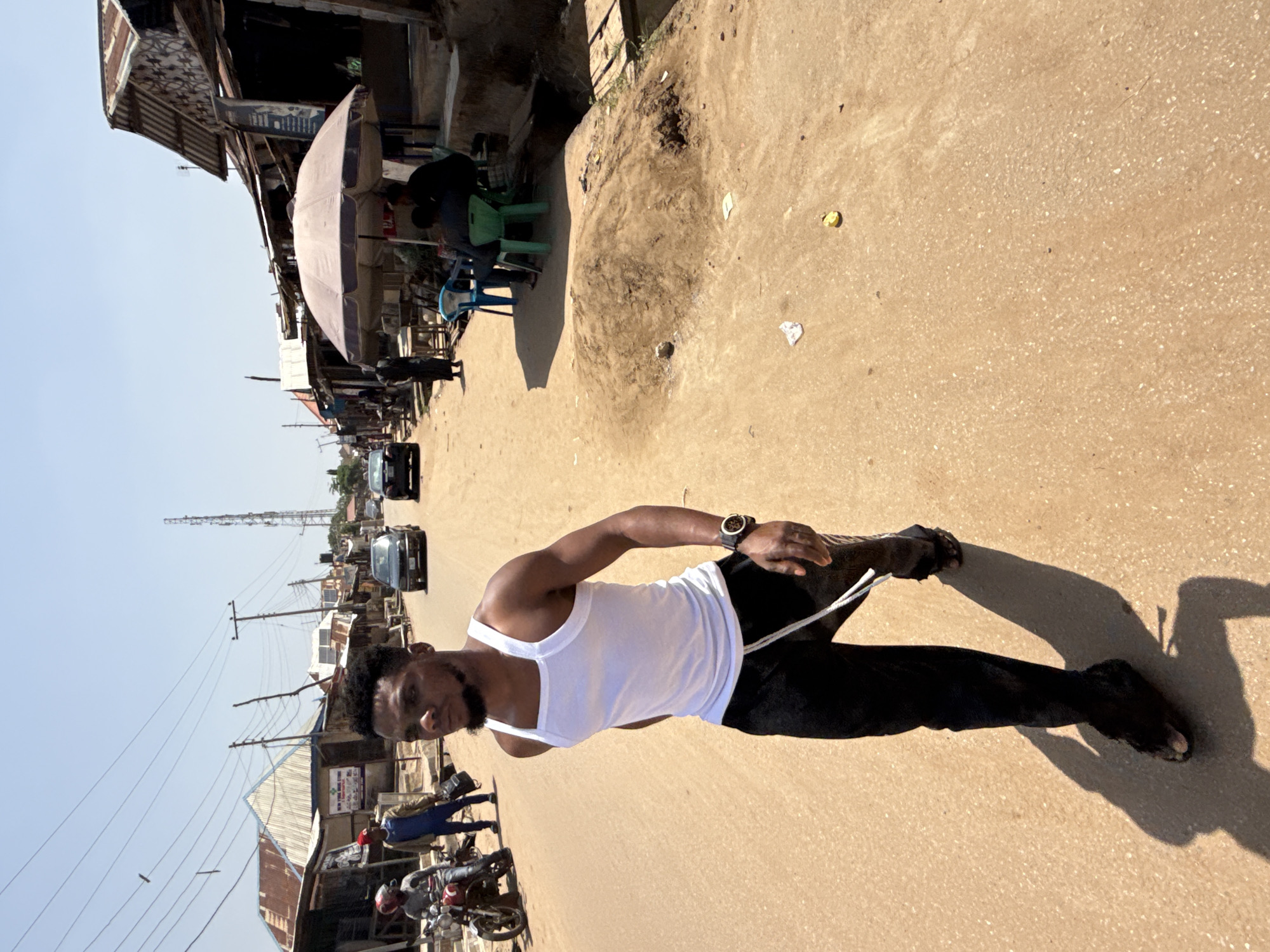 The media file depicts a street scene in Abuja, Nigeria, likely during daytime. A man wearing a white tank top and black trousers is stretching on a sandy, unpaved road. He is looking towards the camera with a neutral expression. He has a beard and short, curly hair. A watch is visible on his left wrist. The setting is a bustling roadside market with various stalls and structures made of corrugated iron and wood. Several motorcycles are parked along the side of the road, and a few cars are visible in the distance. The sky is clear, suggesting sunny weather. In the background, there are power lines and a telecommunications tower. Some debris and litter are scattered on the ground. The overall impression is that of a typical urban environment in Nigeria. No visible text can be identified.