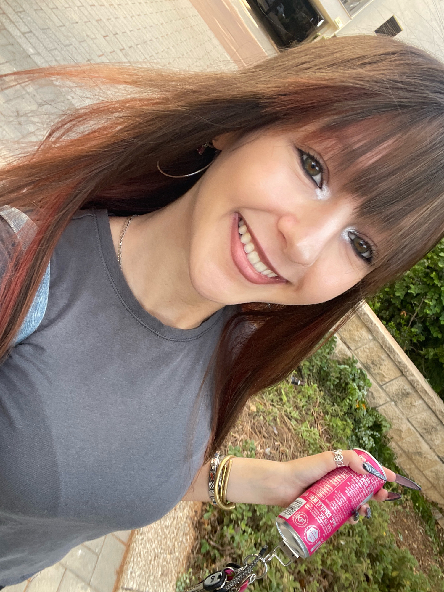 The image shows a young woman with long, brown hair with red highlights, wearing a gray t-shirt and a denim vest. She is holding a pink can of what appears to be a beverage. She is wearing several bracelets and rings, and has long, dark nails. The woman is smiling and looking at the camera.
The background of the image suggests an outdoor urban setting. The ground is paved with light-colored rectangular tiles, indicative of a sidewalk or public walkway. Behind the woman, there is greenery, including bushes and trees, and a stone wall made of stacked, rough-cut stones. The presence of these elements suggests a park, a street with landscaping, or a residential area. The building in the upper right corner, with a window and a tiled facade, further supports an urban environment.
While the image contains elements commonly found in urban areas, there is no specific visual information within the frame that definitively pinpoints the location to the city of Raanana, Israel. The presence of Hebrew text on the pink can is the sole indicator of a possible Israeli context.