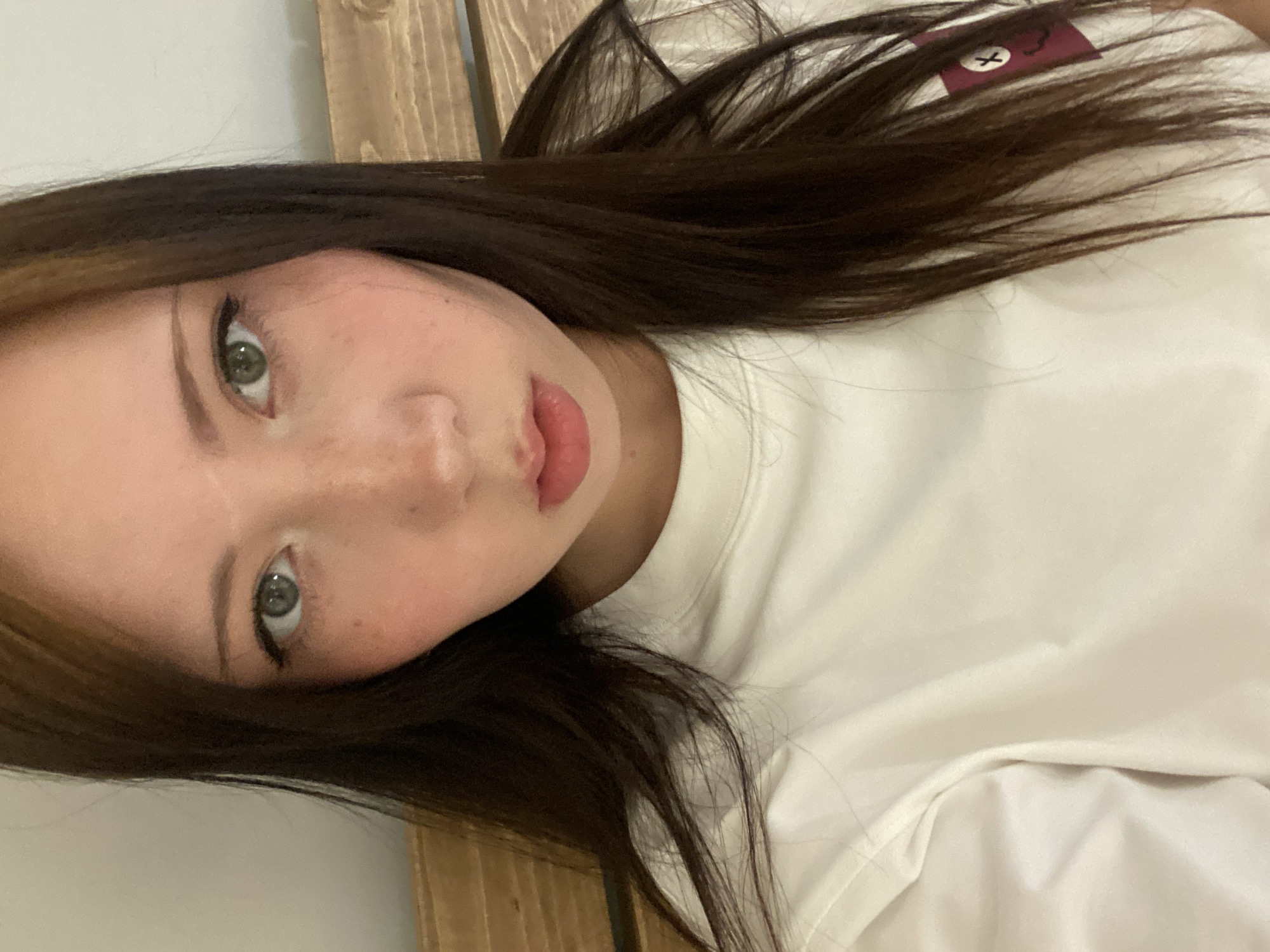 A medium shot depicts a woman with dark hair, fair skin, and green eyes looking into the camera. The woman's face features freckles across the nose and cheeks, with her lips tinted a pale pink. She is wearing a white, long-sleeved shirt. The background consists of what appears to be a wooden headboard. The location and city are unspecified.