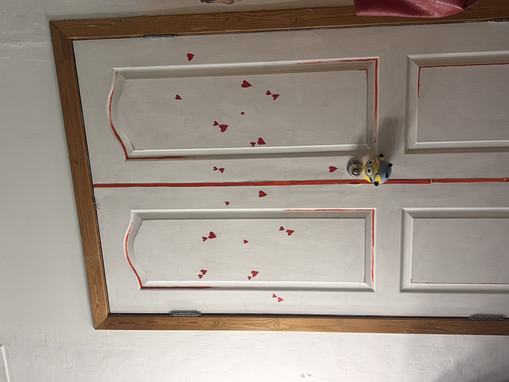 The image displays a white painted wooden door adorned with playful decorations, set within a light-colored room. The door features recessed panels, which are outlined by thin, somewhat uneven hand-painted red lines, suggesting a rustic or DIY aesthetic. Scattered across the white panels are numerous small, red heart-shaped stickers of varying sizes, adding a whimsical touch.
A notable object on the door is a small, yellow and blue Minion character (from the "Despicable Me" franchise) affixed to the door's surface, positioned near a silver-colored knob or latch. This Minion appears to be a decorative element or perhaps a handle cover.
The door itself is framed by a light brown wooden frame with visible grain, contrasting with the white door. A small portion of a red object, possibly a bag or fabric, is visible at the top edge of the door. The wall adjacent to the door is a plain light color, likely white or off-white. No specific activity is occurring; it is a static interior scene. There is no visible text. The overall impression is that of a personalized and decorated door, possibly in a child's room or a home with a playful atmosphere.