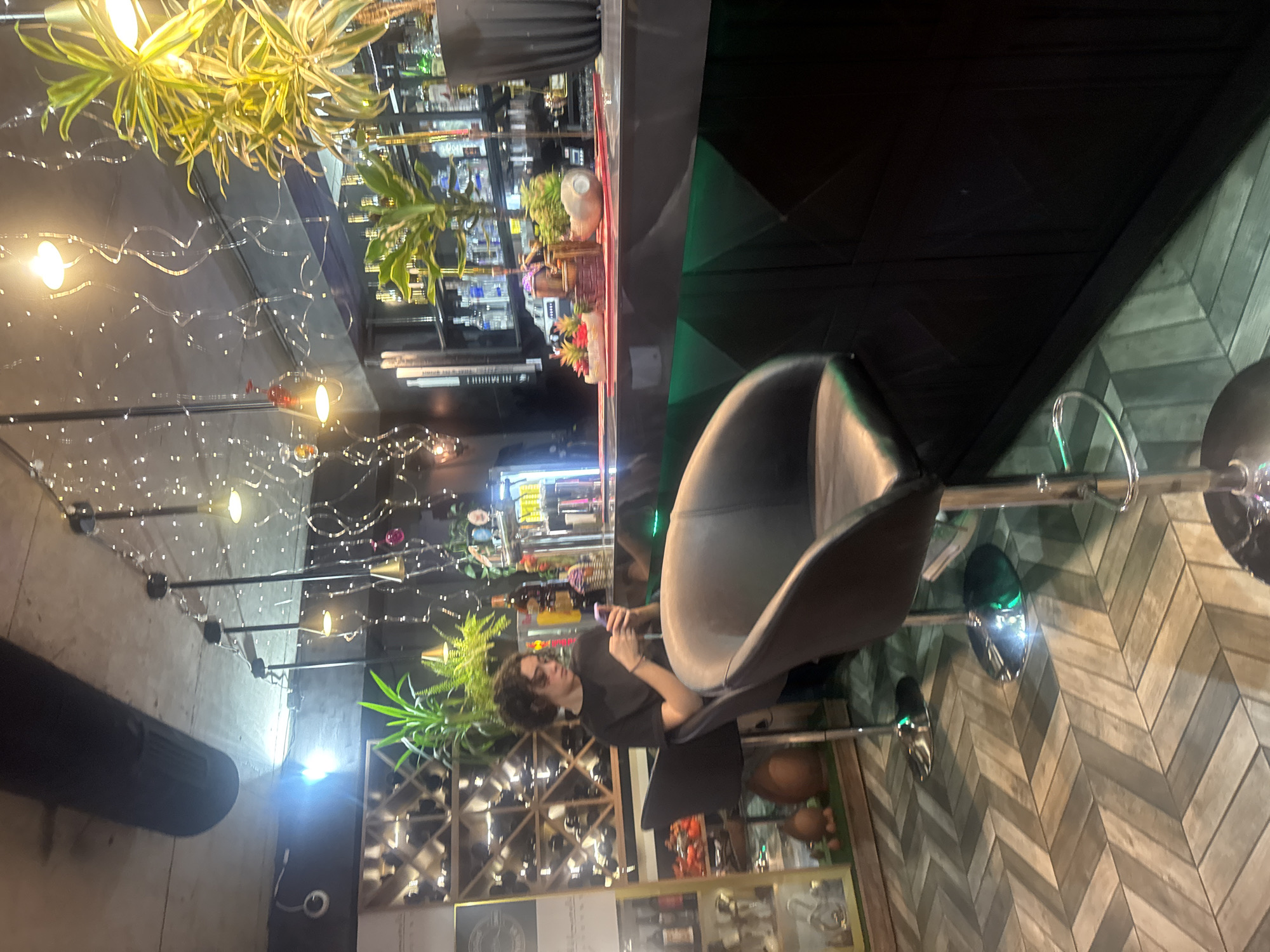A person is seated in a grey bar stool in an indoor setting. The location is likely in Tbilisi, Georgia based on the presence of Georgian signage. The person is wearing a dark shirt and has curly hair. They are looking at a mobile phone. Behind the person is a wine rack and various decorative items. The lighting includes string lights and plant-based lighting fixtures. The background features a bar area with shelves of bottles. The floor has a herringbone pattern.