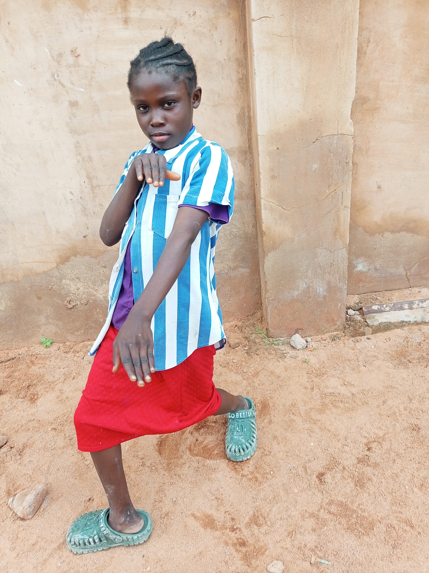 A young girl with braided hair stands in a crouched pose in front of a textured, light brown wall in Tempure, Nigeria. She is wearing a purple shirt underneath a blue and white vertically striped open button-down shirt, and a bright red skirt. Her right leg is bent at the knee, and her left leg is extended slightly behind her. She wears green clogs on both feet. Her left hand is raised, palm outward, as if making a gesture. The ground is covered in light brown sand and small pebbles.