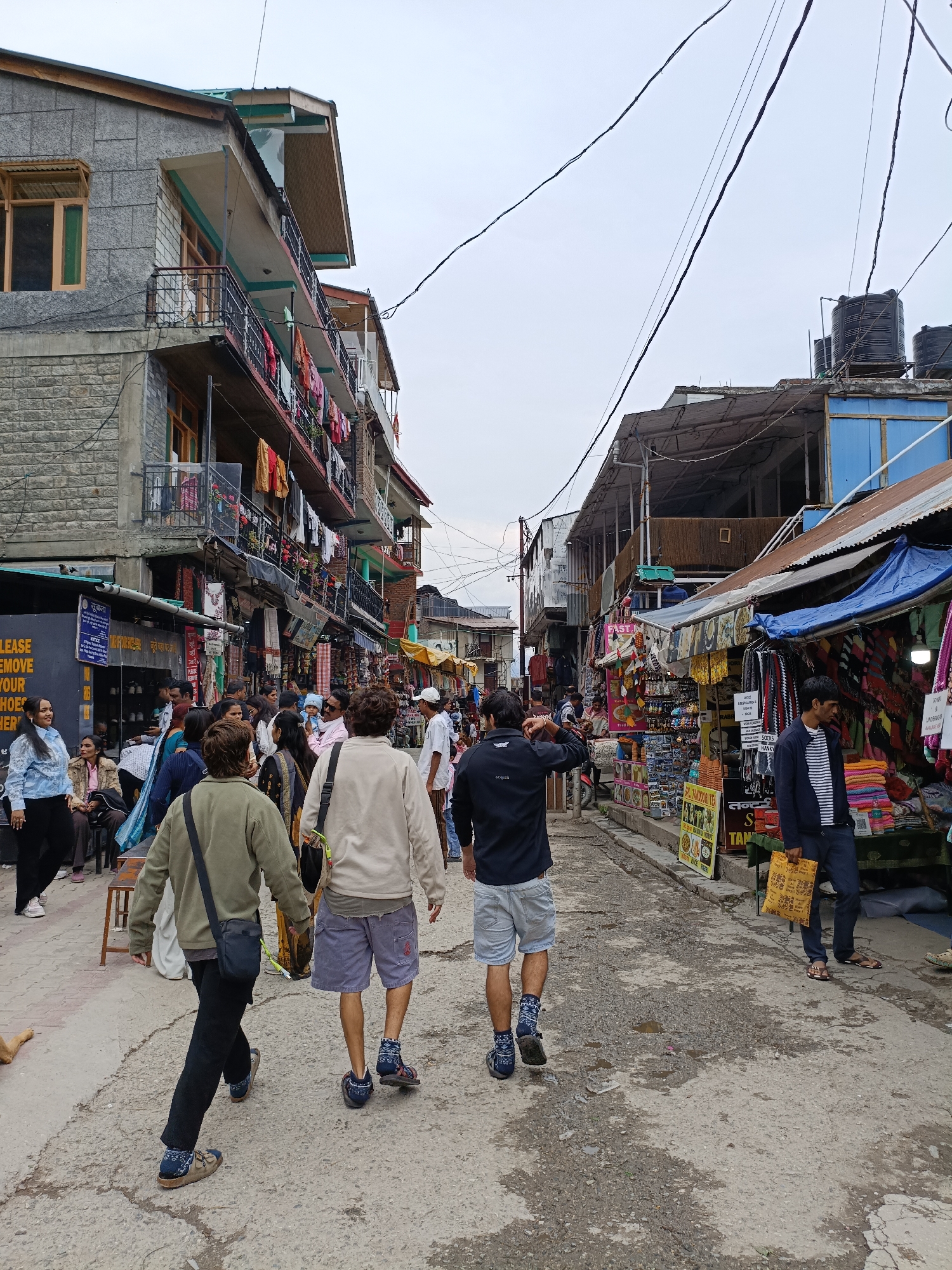 The image captures a bustling street scene in Vashisht, India, with a narrow, paved road lined by multi-story buildings and market stalls. The overcast sky suggests a cloudy day. On the left, a building features stone and concrete construction with balconies adorned with laundry. Market stalls on both sides of the street display a variety of goods, including clothing, bags, and what appear to be food items. Numerous people are walking and browsing, contributing to the lively atmosphere. Some individuals are seen wearing casual clothing, while others are dressed in traditional Indian attire. The ground is uneven and shows signs of wear and tear, with some puddles of water. Overhead, electrical wires crisscross the sky, and water tanks are visible on the rooftops of some buildings. A dog lies on the side of the road in the foreground. The image depicts a vibrant commercial area within a town or village setting.