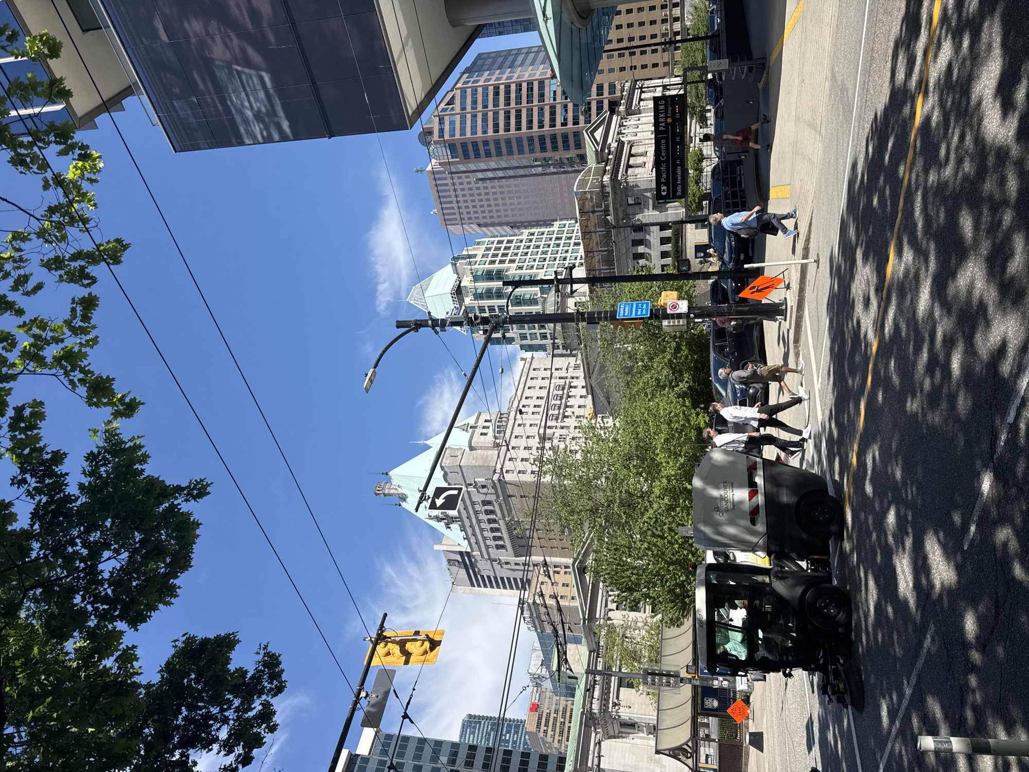 The image captures a street scene in Vancouver, Canada, likely in the downtown core, characterized by a mix of modern and older architecture. Several high-rise buildings are visible against a partly cloudy blue sky. Power lines crisscross the sky above the street. A street sweeper vehicle is parked on the right side of the frame. Pedestrians can be seen crossing the street at a crosswalk. There is a parking information sign. The city of Vancouver logo is visible on the street sweeper. The location is likely near the Pacific Centre, given the presence of its sign. The scene appears to be taken during the day. There are numerous traffic control signs present. The scene is a bustling urban environment with pedestrians and traffic.