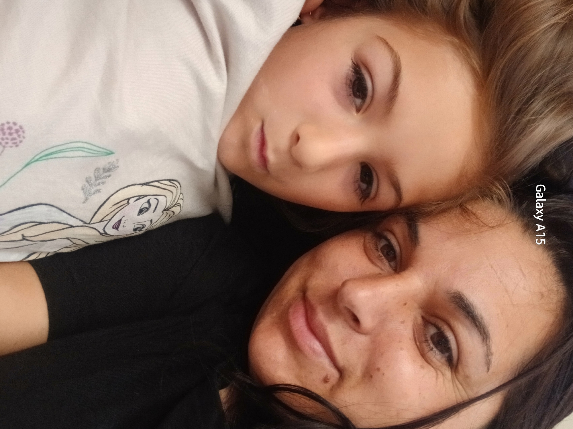 A close-up, top-down shot captures two individuals, appearing to be a mother and daughter, lying down closely together. Both subjects' heads are tilted towards the right, looking towards the camera with soft, calm expressions, suggesting an intimate selfie or portrait.
The younger individual, a child, is positioned in the upper-left part of the frame. She has light brown hair and brown eyes, and her face is smooth. She is wearing a light-colored garment, possibly white or light pink, featuring a partial illustration of Elsa from Disney's "Frozen" along with subtle purple and green floral or leafy designs on the fabric.
The adult woman occupies the lower-right portion of the image. She has darker brown hair and brown eyes, with a slight, gentle smile on her face, conveying warmth and contentment. Her skin shows a few freckles and a mole on her chin. She is dressed in a dark, likely black, top or sweater.
The overall tone of the image is affectionate and peaceful, highlighting the close bond between the two. The lighting is even and natural, consistent with an indoor setting. Along the right edge of the image, the text "Galaxy A15" is visible vertically, indicating the camera used. The location context provided for the image is Obrochishte, Bulgaria.