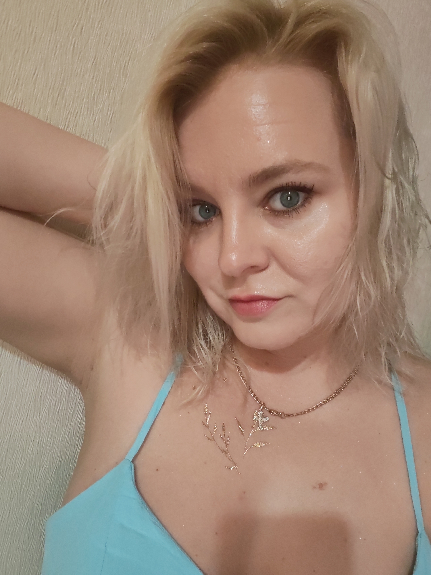 A close-up, head-and-shoulders selfie of a woman with blonde hair and blue eyes. She is wearing a light blue bikini top and a gold chain necklace with a small pendant. The background is a textured wall. The image appears to be taken indoors, likely in a home setting. There is no discernible information about the specific location within Izhevsk, Russia.