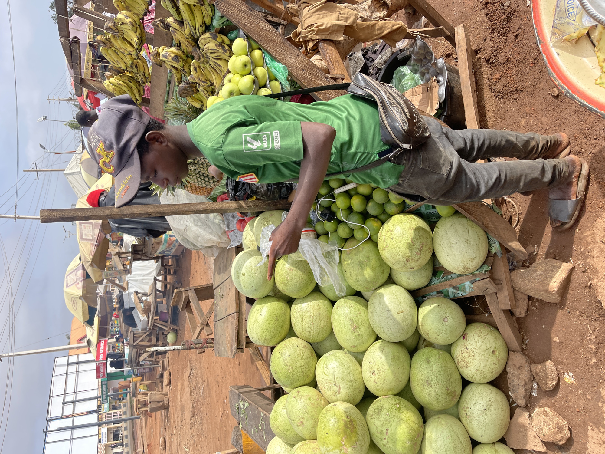 In a bustling street market in Gura Top, Nigeria, a young man wearing a green t-shirt and a gray baseball cap stands at a fruit stand. He is examining a pile of watermelons, holding a clear plastic bag as if preparing to make a selection. Above the watermelons, there are also limes, apples, bananas, and a pineapple for sale. The fruit stand is constructed with wooden supports. The man has a brown sling bag across his body. In the background are other market stalls with umbrellas providing shade. A billboard and power lines are visible in the distance. The ground is covered in reddish-brown soil. The overall scene is one of commerce and activity in an open-air market environment.