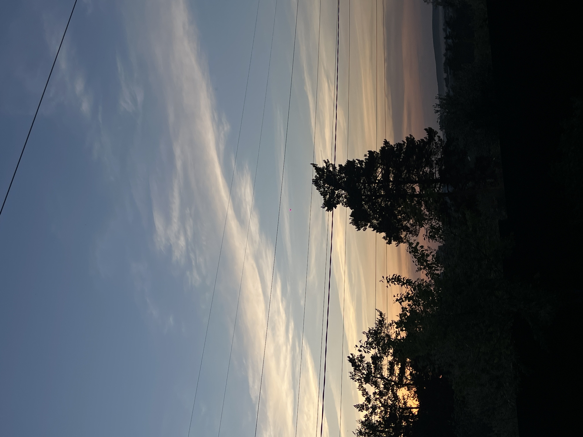 The image depicts a view toward the sunset, likely captured in the city of Chaikovsky, Russia, based on the user's prompt. The sky transitions from a light blue to a warm, orange-yellow hue near the horizon. Wispy clouds stretch across the sky. Overhead power lines run diagonally across the frame. Silhouetted trees and foliage dominate the right side of the image, obscuring some of the sunset. The lower right corner of the frame is dominated by darkness. A small, barely visible red dot is noticeable in the sky, potentially an aircraft beacon or another object. There are no discernible people or specific interactions depicted. The environment appears to be an urban or suburban area bordering a natural environment such as a forest or park.