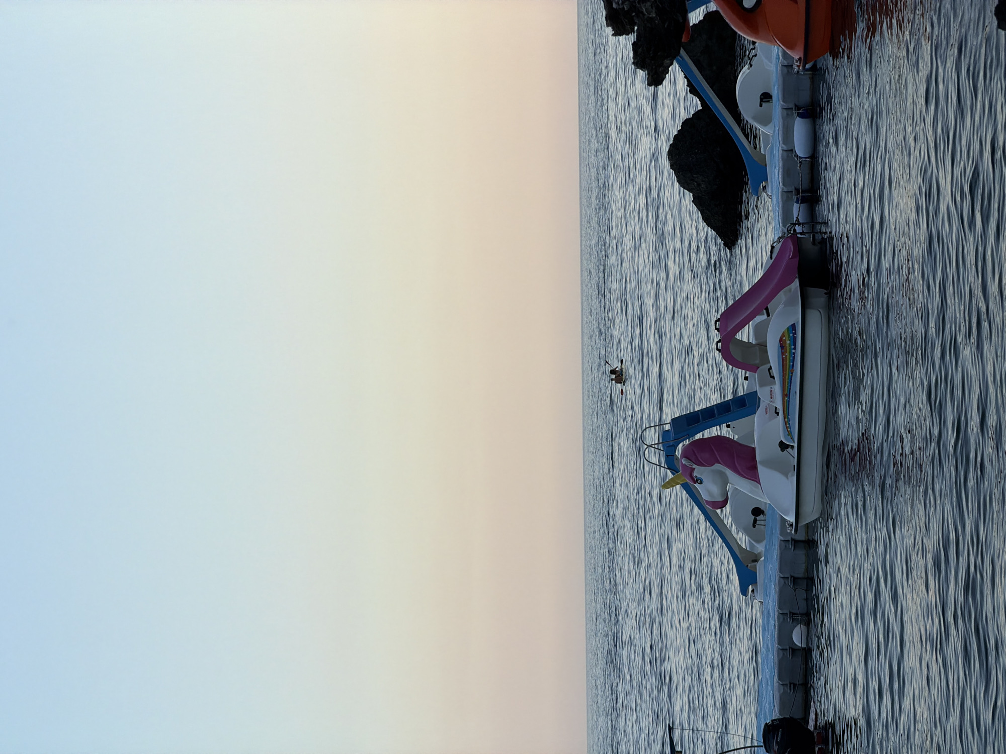 The image depicts a serene coastal scene at dusk, likely in Budva, Montenegro. The sky transitions from a pale blue at the top to soft hues of pink and orange near the horizon, indicating sunset. The sea is calm, with gentle ripples reflecting the twilight sky. In the foreground, a floating dock is moored, supporting several novelty pedal boats. One prominent pedal boat is shaped like a white unicorn with a pink mane and tail, featuring a blue slide. Adjacent to it is another pedal boat with a pink slide. Further to the right, an orange boat with a large eye graphic is visible. Two individuals are seen in the distance on a paddleboard, enjoying the water. Rocky outcrops are present along the coastline on the right side of the image. The overall atmosphere is peaceful and tranquil, capturing a moment of relaxation by the Adriatic Sea.