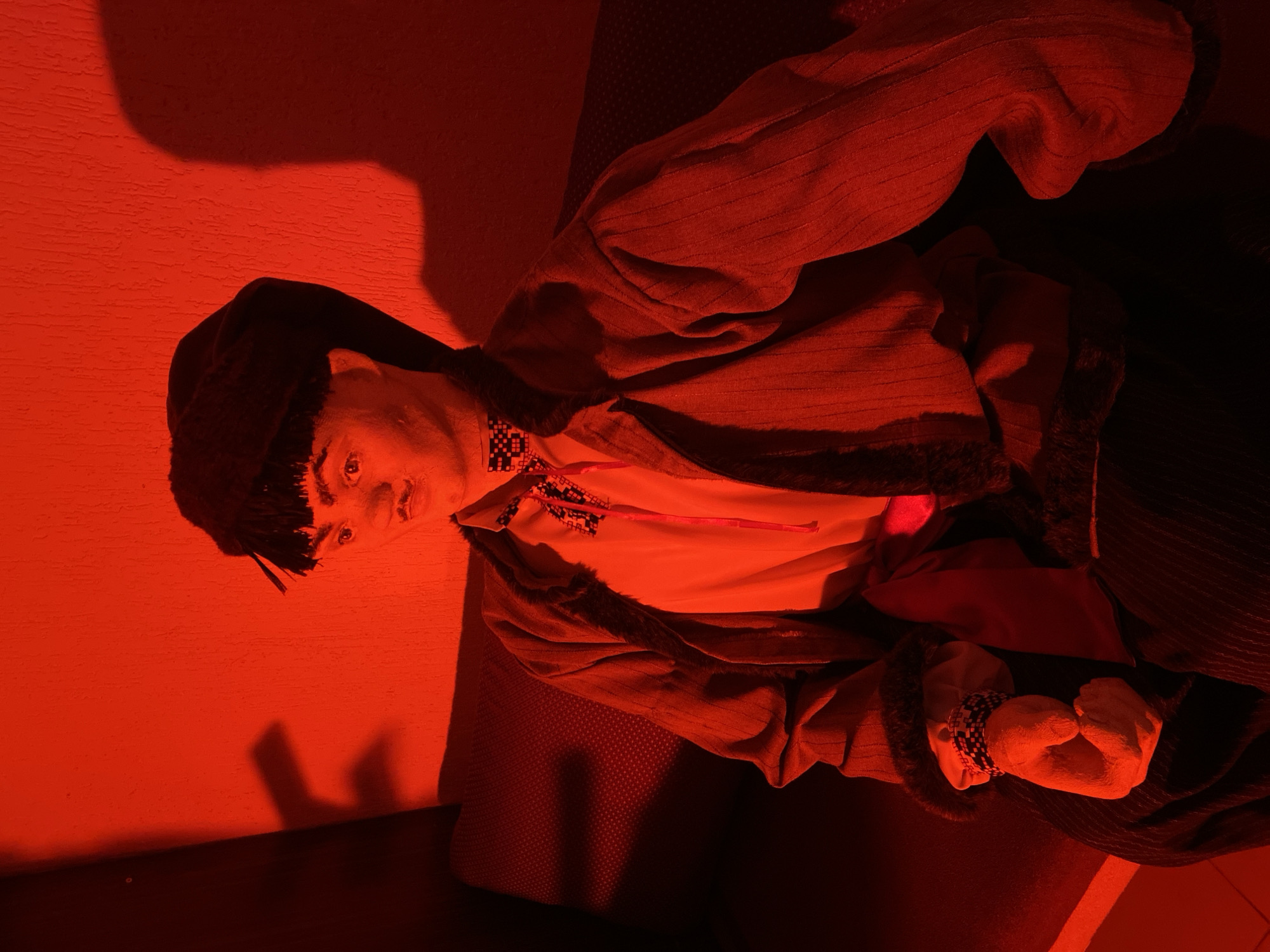A mannequin dressed in traditional Russian clothing is displayed indoors. The figure is wearing a dark fur hat, a white shirt with embroidered details, and a long jacket with fur trim. The scene is bathed in a red light, obscuring details. The mannequin is located indoors, likely in Chaikovsky, Russia, based on the origin of the image.