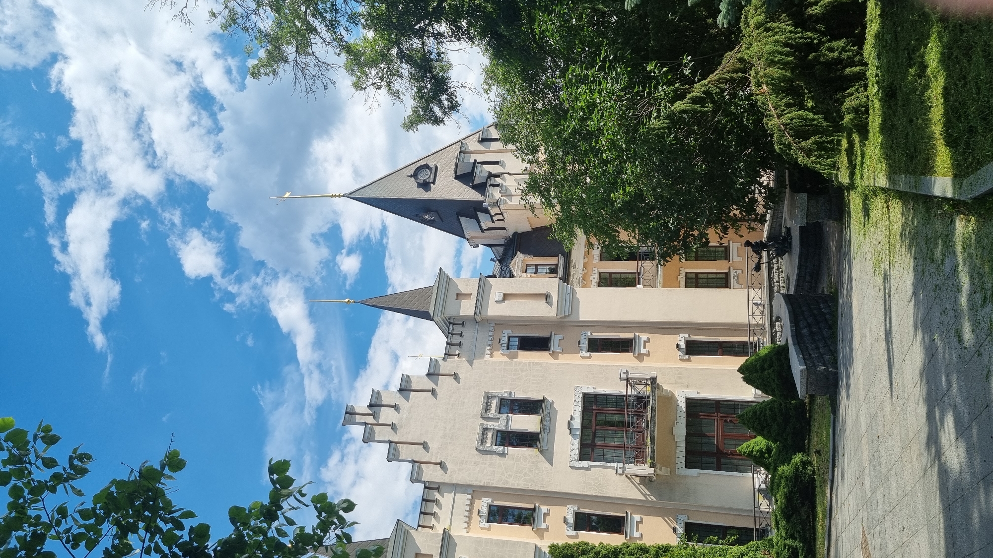 This media file shows a view of a unique, fairytale-like building with multiple spires and ornate architectural details. The building appears to be made of light-colored stone or stucco and has a cream-colored facade with a textured finish. Several windows are visible, some with dark frames and what appear to be intricate metalwork on balconies. The spires are dark gray or black, tapering to sharp points, with decorative weather vanes.
The building is surrounded by lush greenery, including mature trees with dense foliage and neatly manicured evergreen bushes. In the foreground, there is a paved walkway or patio made of stone tiles, with some grass areas adjacent to it. Part of the walkway is in shadow, suggesting it is either morning or late afternoon.
The sky is a vibrant blue, dotted with fluffy white clouds, indicating fair weather. The lighting suggests it is daytime, likely a sunny day. There are no people visible in the frame, and the focus is entirely on the architecture and its natural surroundings. The overall impression is one of a whimsical and picturesque location, possibly a historical landmark, a private residence with unique design, or a place of cultural significance in Kyiv, Ukraine. The architectural style is reminiscent of Gothic or romanticized medieval designs.