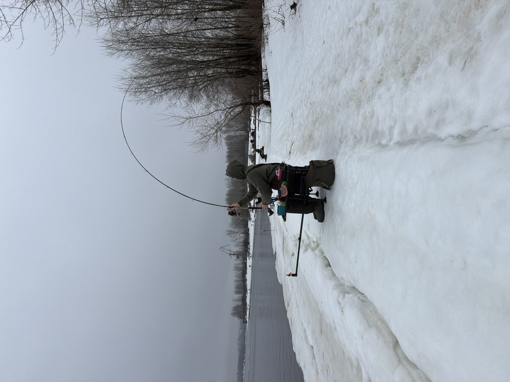 A person wearing a green hooded jacket and dark trousers is ice fishing on a snowy bank along a body of water. The person is holding a fishing rod bent in an arc. The person is seated on a platform. Fishing equipment is visible near the person. The location appears to be Chaikovsky, Russia, based on recognizable elements in the background. The weather is overcast, and there are bare trees behind the fisherman. In the distance, there's a glimpse of industrial structures. The foreground shows layers of snow and ice.
