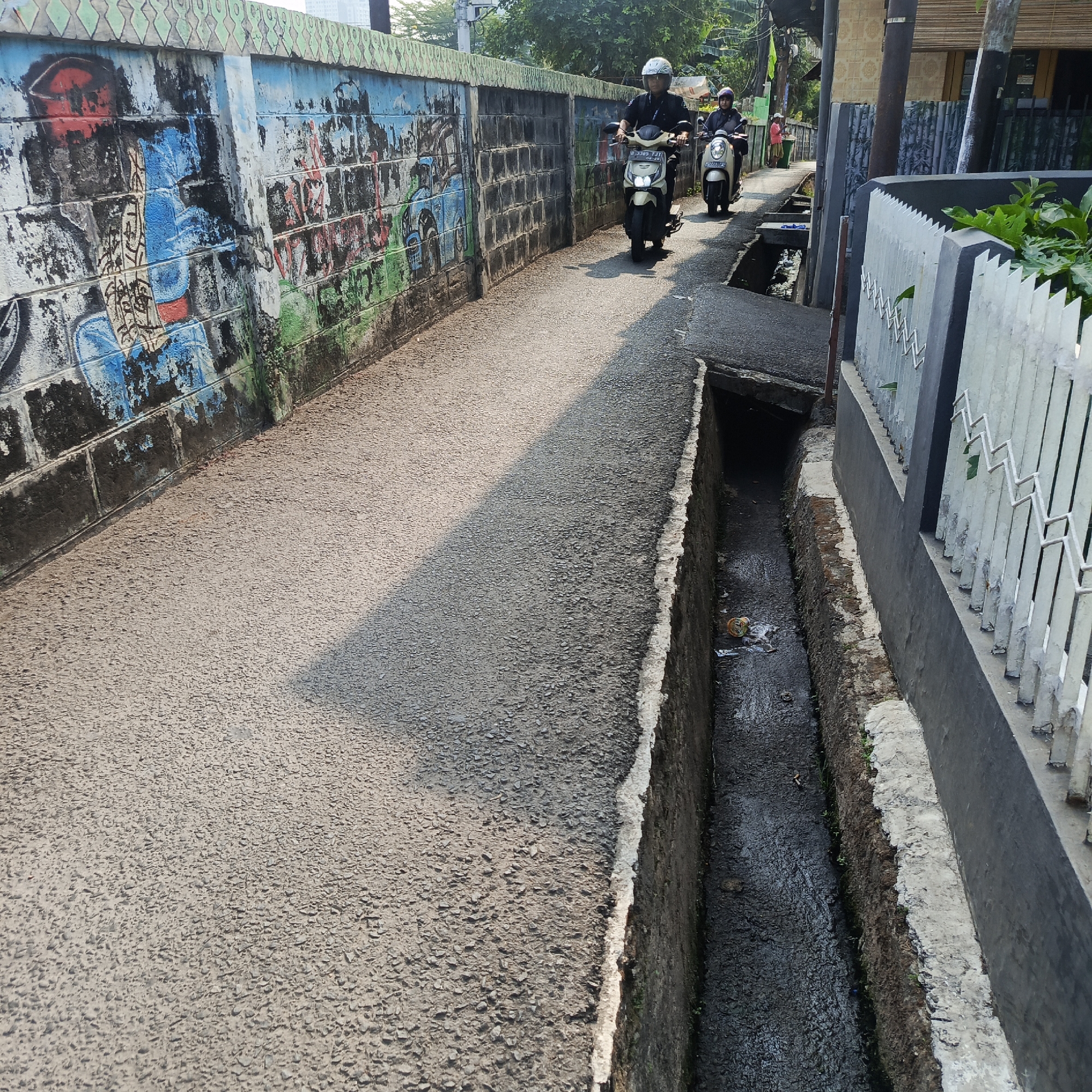 Two individuals on motorbikes are riding down a narrow, paved alley in South Jakarta, Indonesia. The alley is flanked on the left by a weathered brick wall covered in graffiti, featuring colorful but faded artwork. On the right, a concrete ditch runs alongside the road, containing dark, murky water and some debris. Beyond the ditch, a white metal fence and a building are visible. The lighting suggests it is daytime, with shadows cast across the pavement from the buildings and a tree. The overall scene depicts a typical urban environment, likely a residential area, with elements of everyday life and street art.
