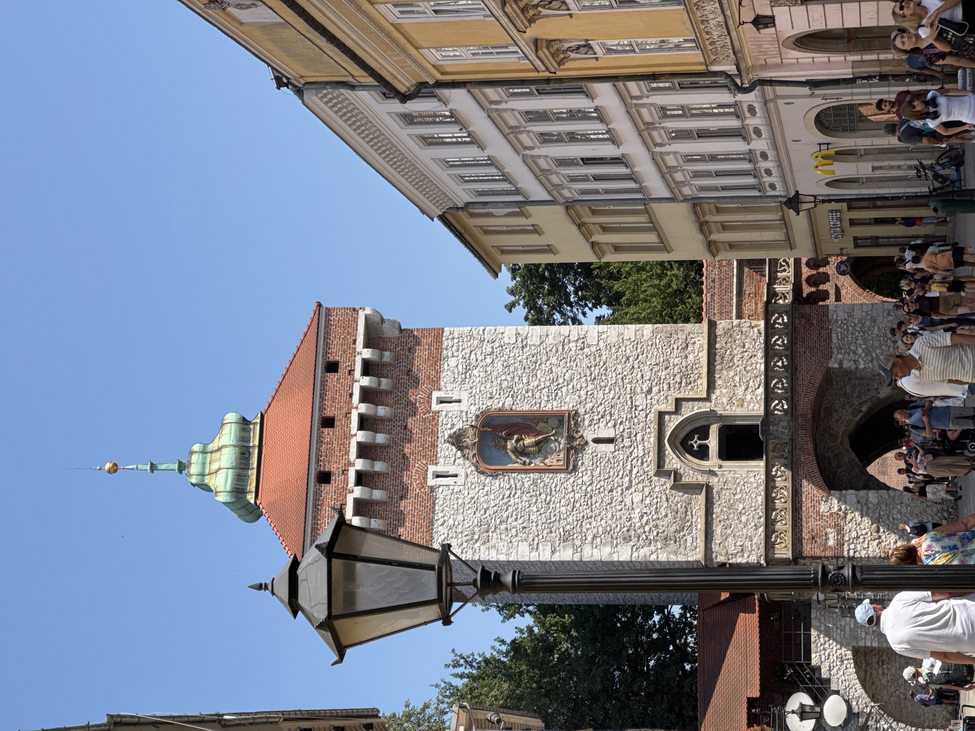 This image shows a street view in Krakow, Poland, specifically depicting the Florian Gate (Brama Floriańska). The dominant structure is the historic Florian Gate, a medieval tower made of stone and brick. It features a prominent pitched red tile roof and a green, onion-shaped dome topped with a golden finial. A decorative framed artwork depicting a warrior is mounted on the stone facade of the gate, above a gothic-arched window. A large, black, ornate lamppost stands in the foreground, obscuring part of the gate. To the right of the gate are several multi-story buildings with a variety of architectural styles, featuring arched doorways and numerous windows. These buildings appear to house shops, including a McDonald's and a "COSTA COFFEE". A street lined with people is visible, with individuals walking and standing. The sky is clear and blue, indicating a sunny day. Trees are visible behind the gate, adding greenery to the scene. The overall atmosphere is that of a busy, historic urban environment.