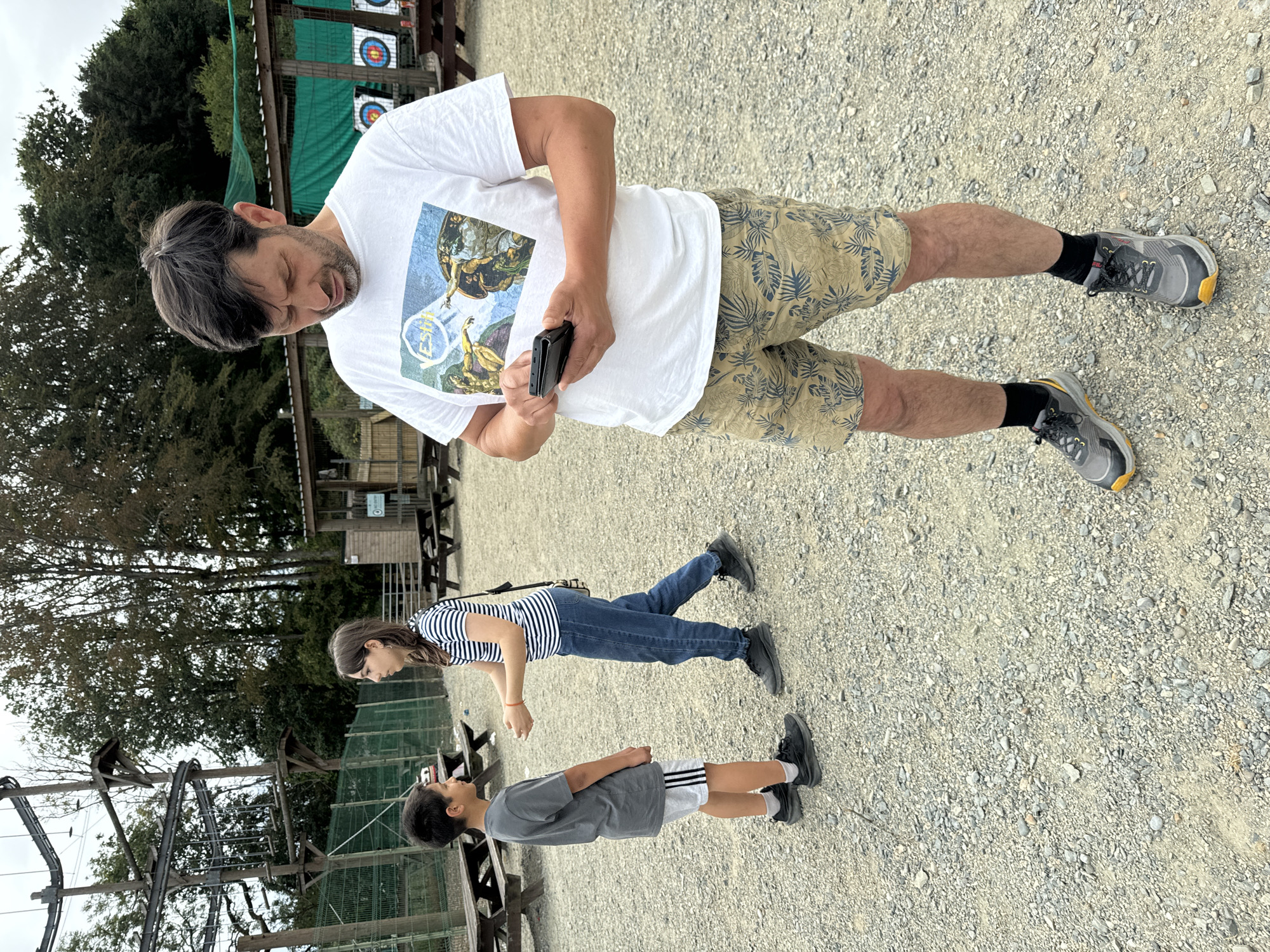 In Courtown, Ireland, on a cloudy day, a man stands holding a black smartphone in his hands, looking down at it. He is wearing a white t-shirt with a graphic print and patterned shorts, black socks and grey and yellow athletic shoes. In the background, a boy in a grey t-shirt and white shorts is facing a girl in a striped shirt and blue jeans, with their hands gesturing towards each other. They appear to be in an outdoor activity center, with wooden benches and picnic tables scattered around. In the distance, a high ropes course structure is visible, along with archery targets. The ground is covered in gravel.