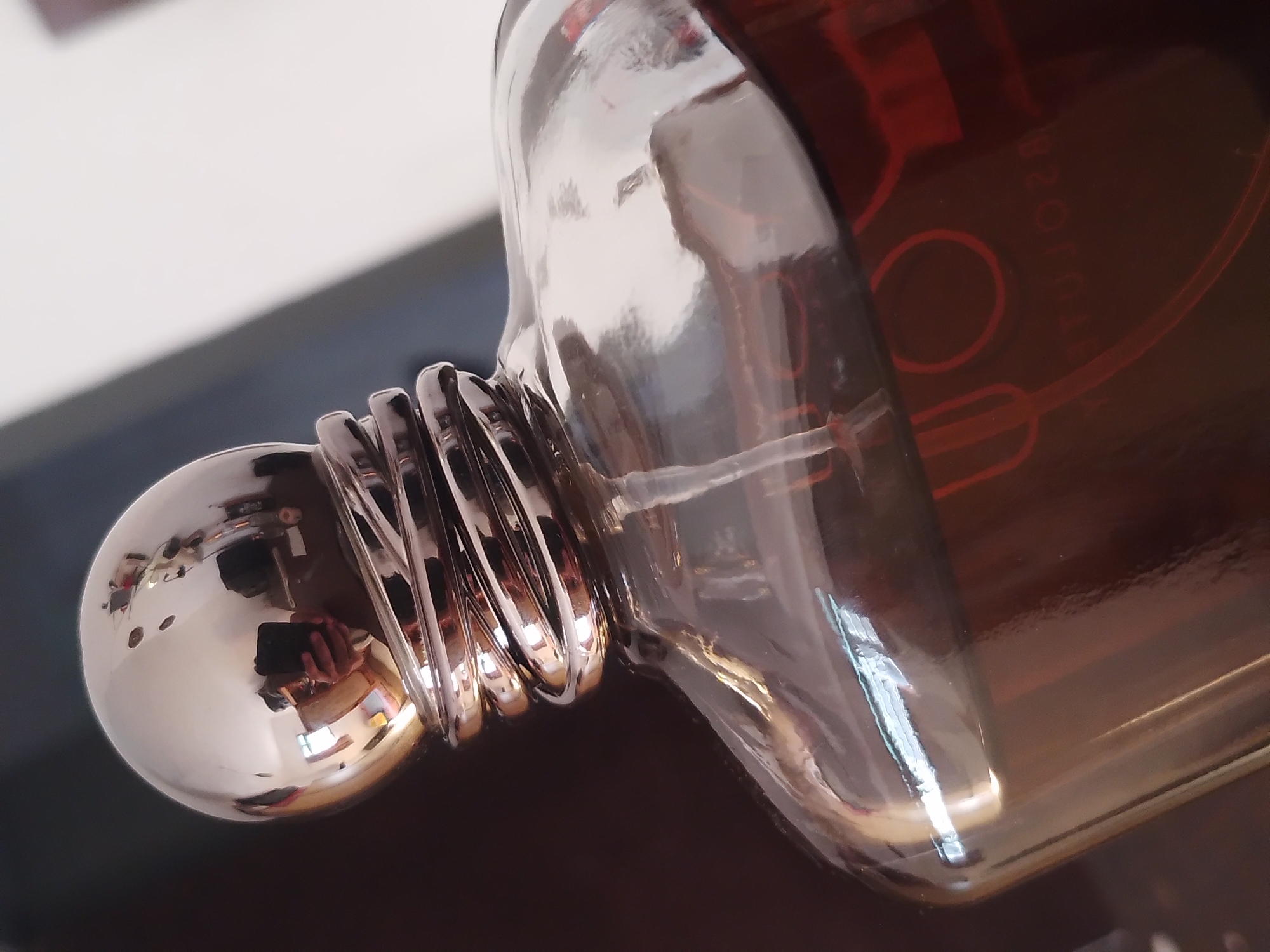 A close-up, eye-level shot focuses on the top portion of a clear glass perfume bottle filled with a dark amber liquid. The bottle has a distinctive metallic, coiled neck that transitions into a rounded, silver cap. The reflective surfaces of the cap and bottle show distorted reflections of the surrounding environment, including what appears to be a person holding a phone. The background is dark and out of focus, with a lighter, out-of-focus area on the right. Red lettering is faintly visible through the glass on the front of the bottle. The image was taken in Batu, Indonesia, and the scene depicts an indoor setting, likely a vanity or table, with artificial lighting contributing to the reflections.