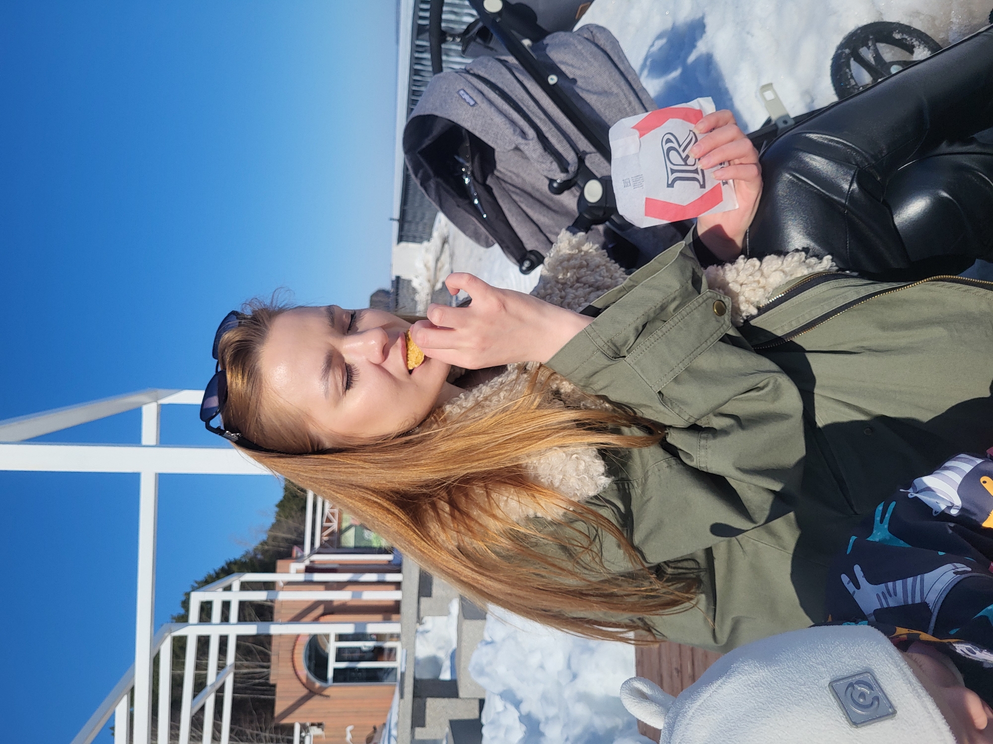 Here is a description of the image:
The image shows a person with long, red hair eating something yellow. They are wearing a green jacket with a fur-lined collar and black pants, possibly leather. They are holding a white package with black lettering, possibly from a restaurant.
Behind the person, there is a baby stroller and snow on the ground. A white metal railing is also visible. In the background, there is a building and trees under a bright blue sky.
Based on the architectural style and the presence of snow, the location is likely in Russia, possibly Chaikovsky. The scene appears to be outdoors on a sunny winter day.