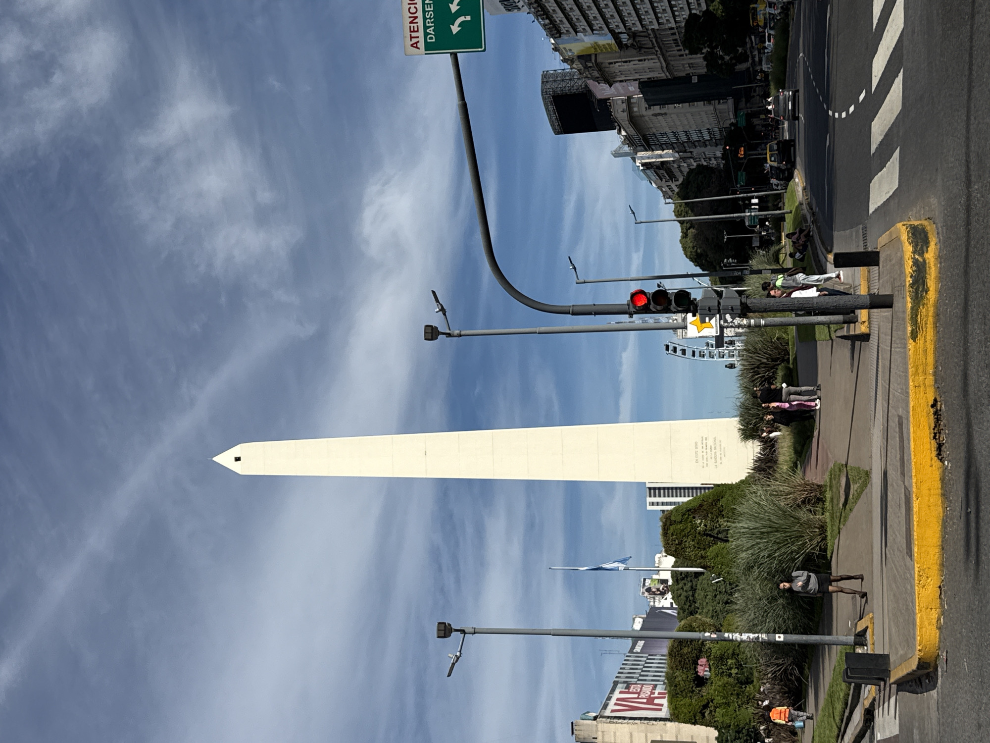 The image captures the Obelisco de Buenos Aires, a national historic monument in Plaza de la República, Buenos Aires, Argentina. The obelisk, a tall, white rectangular pillar, dominates the left side of the frame, extending upwards towards a cloudy blue sky. The surrounding environment includes buildings of varying heights, trees, and landscaped greenery. Traffic lights are visible, indicating active city infrastructure. People are seen walking and standing on the sidewalks, suggesting a public space with pedestrian activity. The angle and perspective suggest the photo was taken at street level, close to the obelisk. There's a yellow curb bordering the sidewalk, and a pedestrian crossing is also visible, further emphasizing the urban street scene. A road sign is visible in the upper left corner. Overall, it's a vibrant city scene featuring an iconic landmark.