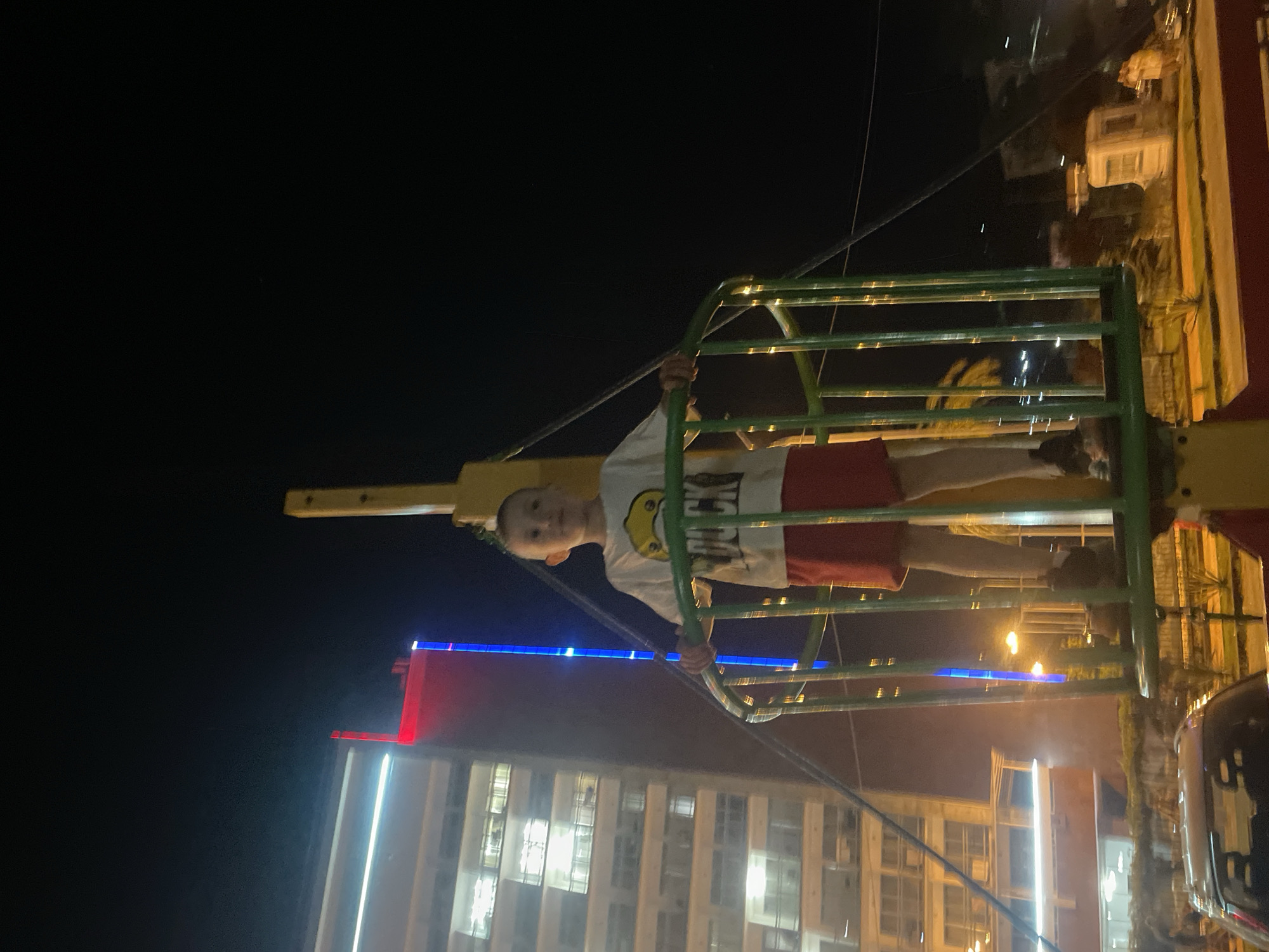 A young boy stands in a green gondola at night, holding onto the railing. He is wearing a white t-shirt with a cartoon duck graphic and red shorts. The gondola is part of a larger amusement park ride, possibly a Ferris wheel, as indicated by the yellow mast behind him and the cables extending from the structure. In the background, a tall, modern building with illuminated windows is visible, along with red and blue lights on its facade. The scene appears to be in a city, with streetlights and other buildings faintly visible in the distance. The overall impression is of a lively evening amusement in an urban setting. The location is Gagra, Abkhazia.