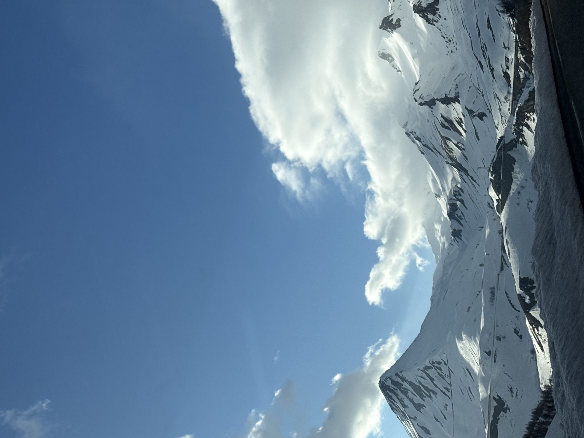 The image shows a view of snow-covered mountains under a partly cloudy blue sky. The mountain range dominates the right side of the frame, with the snow highlighted by sunlight. Clouds are interspersed among the peaks and in the sky. The location is Le Monêtier-les-Bains, France.
