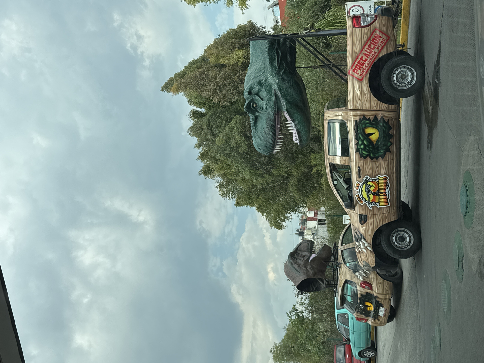 The image shows a roadside attraction in San Bernardino Tlaxcalancingo, Mexico. Two pickup trucks, possibly Ford Rangers, are parked side-by-side, decorated with a "Jurassic Park/Dino-Sound" theme. The trucks have a faux-wood grain exterior with dinosaur-related decals, including an eye and claw marks. A large, green Tyrannosaurus Rex head is mounted on a metal frame above one of the trucks. A sign in Spanish on the back of the truck reads "Precaucion Dinosauros Vivos." The background includes green trees, an overcast sky, buildings, and other parked vehicles.
