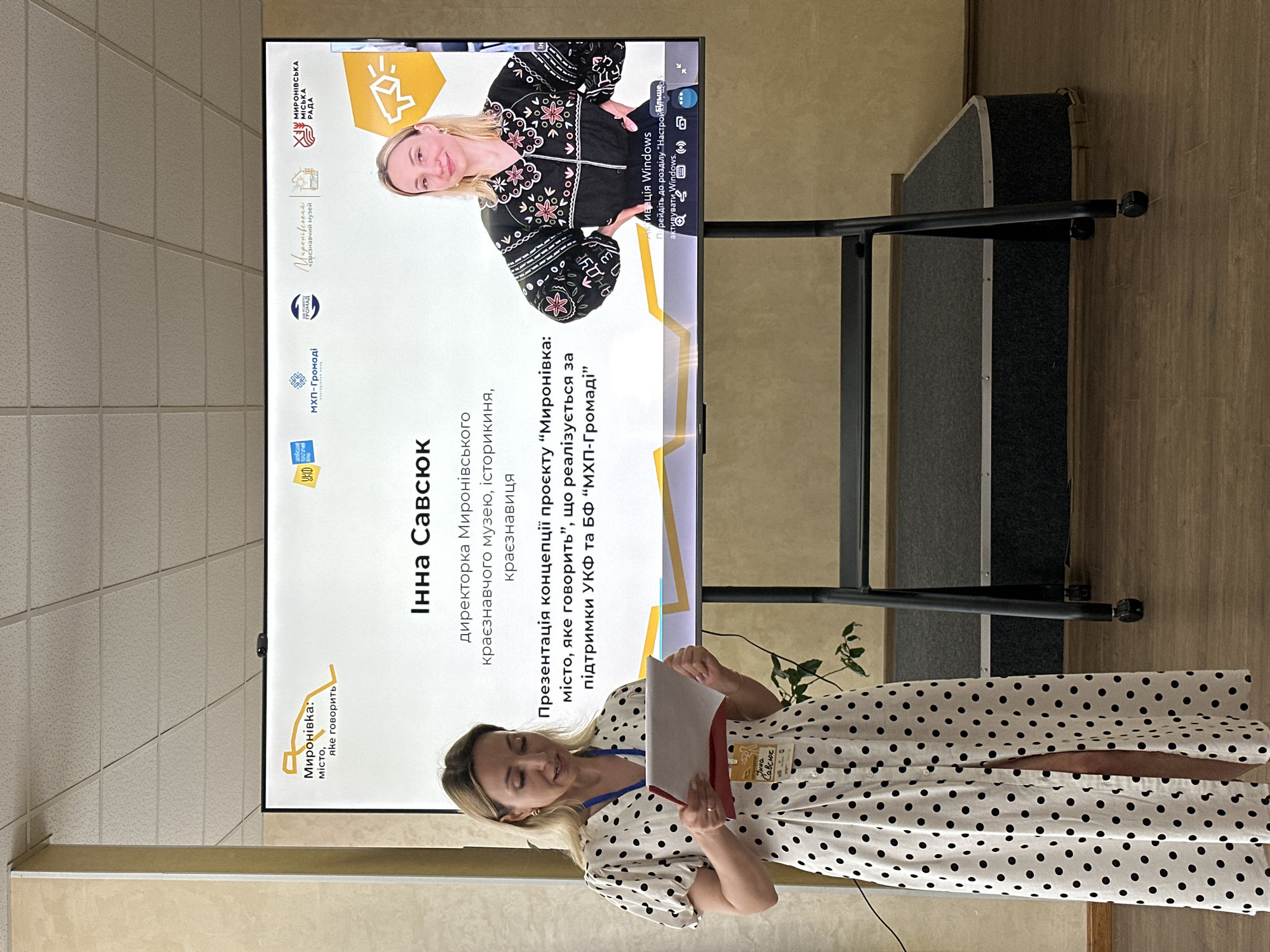 This media file captures a presentation taking place indoors, likely in a community hall or conference room. The primary subject is a woman, identified by the text on the screen as Inna Savsyuk, the director of the Myronivka Regional Museum. She is standing in front of a large screen displaying presentation slides, and she is holding a document and appears to be speaking.
The screen prominently features text in Ukrainian, indicating the presentation is about a project titled "Myronivka: The City That Speaks." It also mentions that the project is being implemented with the support of the Ukrainian Cultural Foundation and the MHP-Community Foundation. The screen also displays logos of supporting organizations, including "Myronivka City Council" and the "Myronivka Regional Museum." A photograph of Inna Savsyuk is visible on the top right of the screen.
Inna Savsyuk is wearing a black and white polka dot dress and a lanyard with an identification badge. The overall atmosphere appears professional and informative, with the woman engaged in delivering her presentation. The lighting suggests it is daytime, and the indoor setting implies no specific weather conditions are relevant. The scene is set in Myronivka, Ukraine, as indicated by the text on the screen.