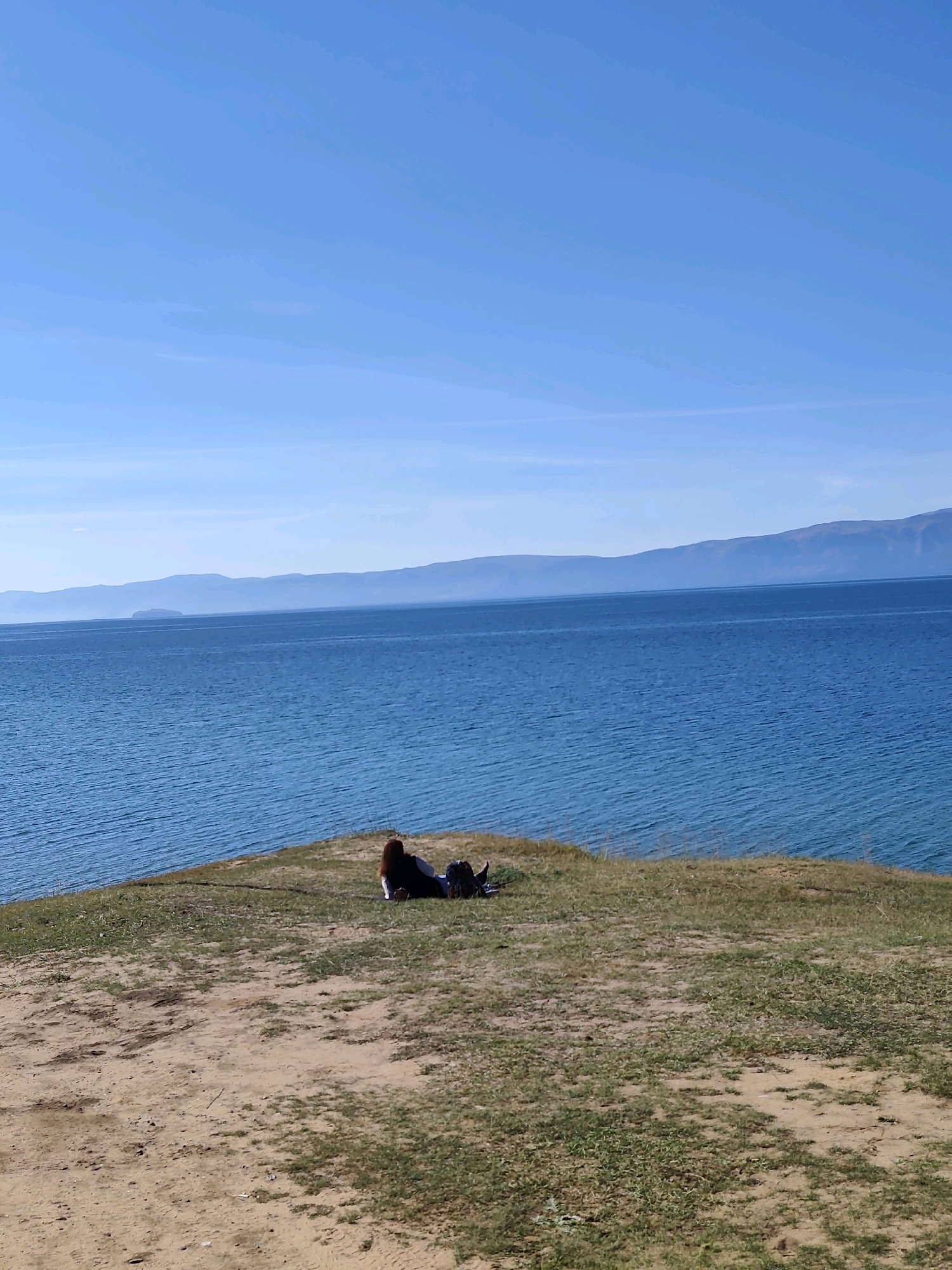 In Kharantsy, Russia, a person is lying on their side on a grassy knoll overlooking a large body of water. The person is wearing dark clothing and has reddish-brown hair. They are positioned on the right side of the frame. The water, a vibrant blue, stretches to the horizon where it meets hazy, distant mountains under a clear blue sky. The foreground consists of a patch of dry, sandy earth interspersed with green grass. The overall scene depicts a serene, natural landscape.