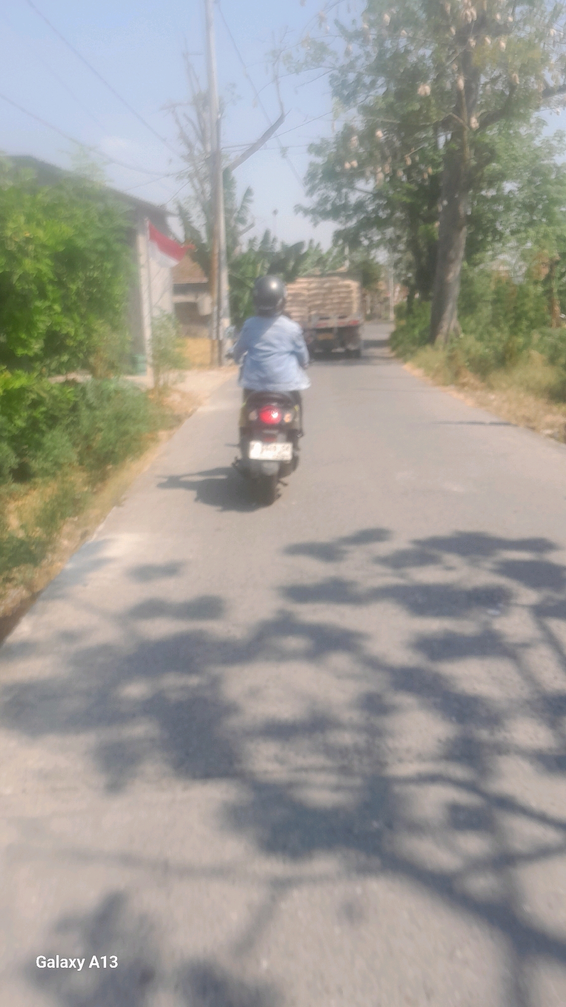 The image shows a person riding a scooter on a road in Kudus, Indonesia. The scooter is black with a license plate that appears to read "K 4934 AB". The rider is wearing a blue shirt and a black helmet. In the background, there is a truck loaded with what appears to be hay or straw, and on the left side of the road, there is a building with a red and white Indonesian flag. The scene is lit by sunlight, casting shadows of trees on the road. The image is captured from behind the scooter.