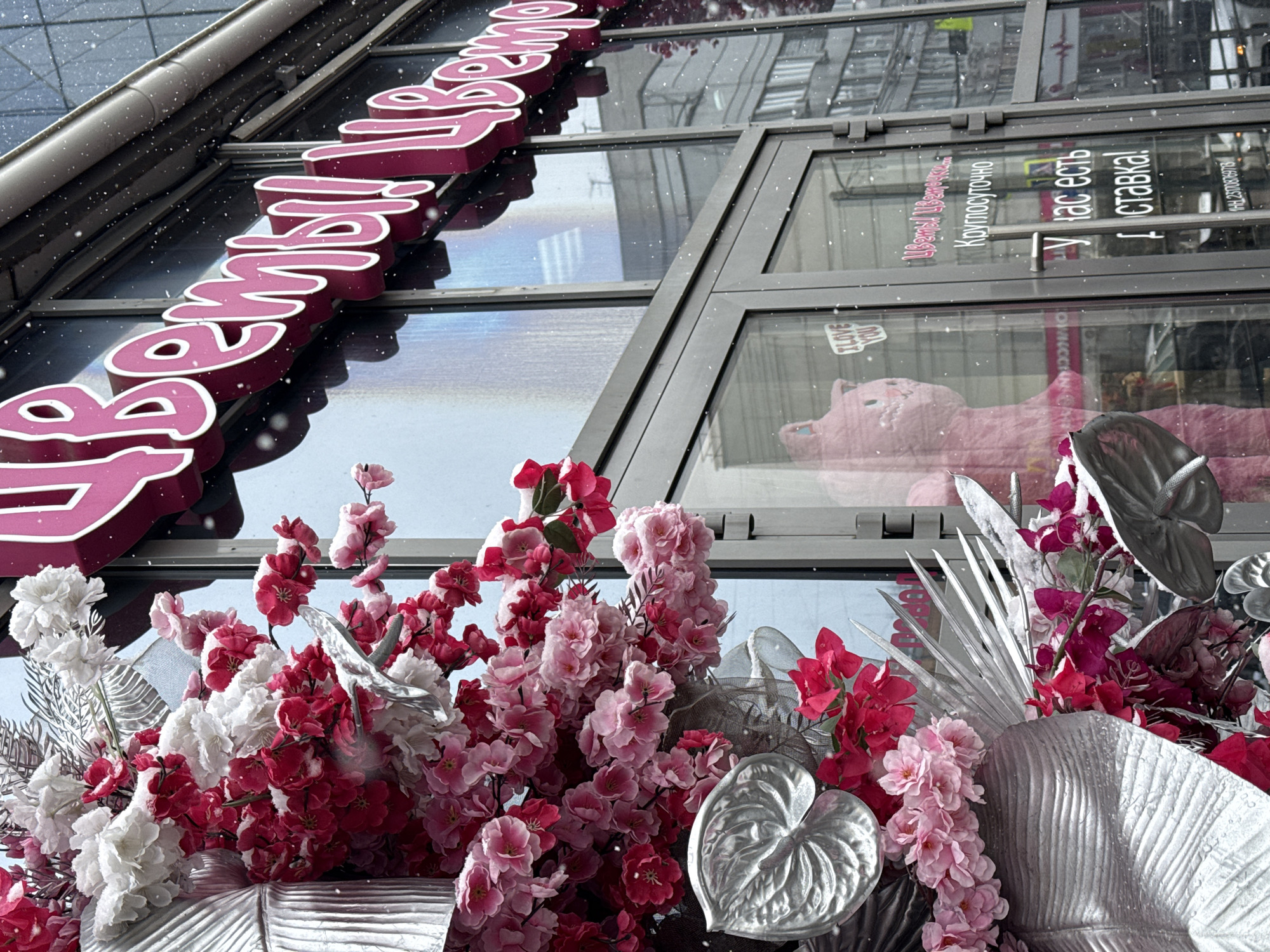 This upward-angled image captures the vibrantly decorated storefront of a building, likely a flower shop, during a light snowfall in Krasnoyarsk, Russia. The foreground features a rich display of artificial flowers in shades of dark red, various pinks, and white, mixed with striking silver foliage resembling palm leaves and anthuriums. Small white snowflakes are visible, creating a wintery atmosphere.
Above the floral arrangement are large glass windows that reflect the urban surroundings. Through the glass, a sizable pink plush toy, possibly a bear, can be seen. A prominent pink sign with white trim is mounted on the building's facade, displaying the word "Фламинго" (Flamingo) in Cyrillic script.
Text printed vertically on the glass windows informs customers: "Цветы" (Flowers), "Круглосуточно" (24 hours), "У нас есть" (We have), and "Доставка!" (Delivery!). A small "I LOVE YOU" sticker is also visible on a lower window pane, and a Taxpayer Identification Number (ИНН 23070269781) is listed in small print on the window frame. The scene portrays a bustling floral business operating with extended hours amidst snowy weather.