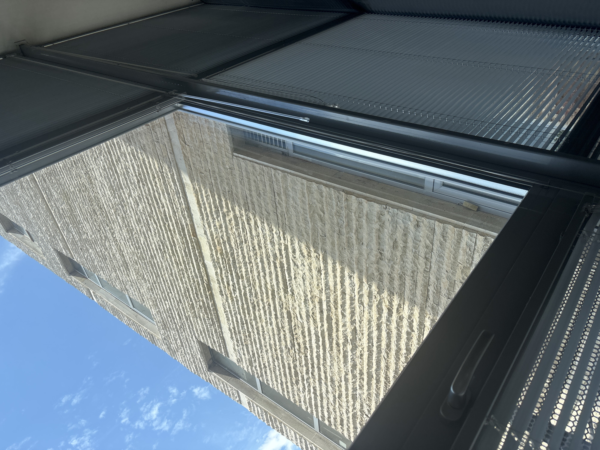 This media file captures an upward-looking view from inside a modern building, showing an exterior scene under a bright sky. The perspective is heavily tilted.
In the foreground, a dark gray, sleek window or skylight frame is visible, including a dark handle on the right side. Above the glass, there's a sophisticated shading system composed of dark, horizontal slats or louvers, some of which appear to be perforated or have a mesh-like texture. Wires or thin rods are integrated into this system, suggesting it is adjustable or retractable.
Through the window, the prominent subject is the exterior wall of an adjacent building. This wall is constructed from a light-colored, possibly beige or off-white, material with a distinctive, deeply textured surface characterized by uniform vertical grooves. Several rectangular windows with dark frames are visible on this building facade.
Above the building wall, a clear, bright blue sky with sparse, wispy white clouds dominates the background, indicating fair weather and daytime. The bright sunlight casts some shadows, highlighting the texture of the stone wall.
The scene depicts a modern architectural environment in Blagoevgrad, Bulgaria, under sunny conditions. There is no discernible activity or visible text.